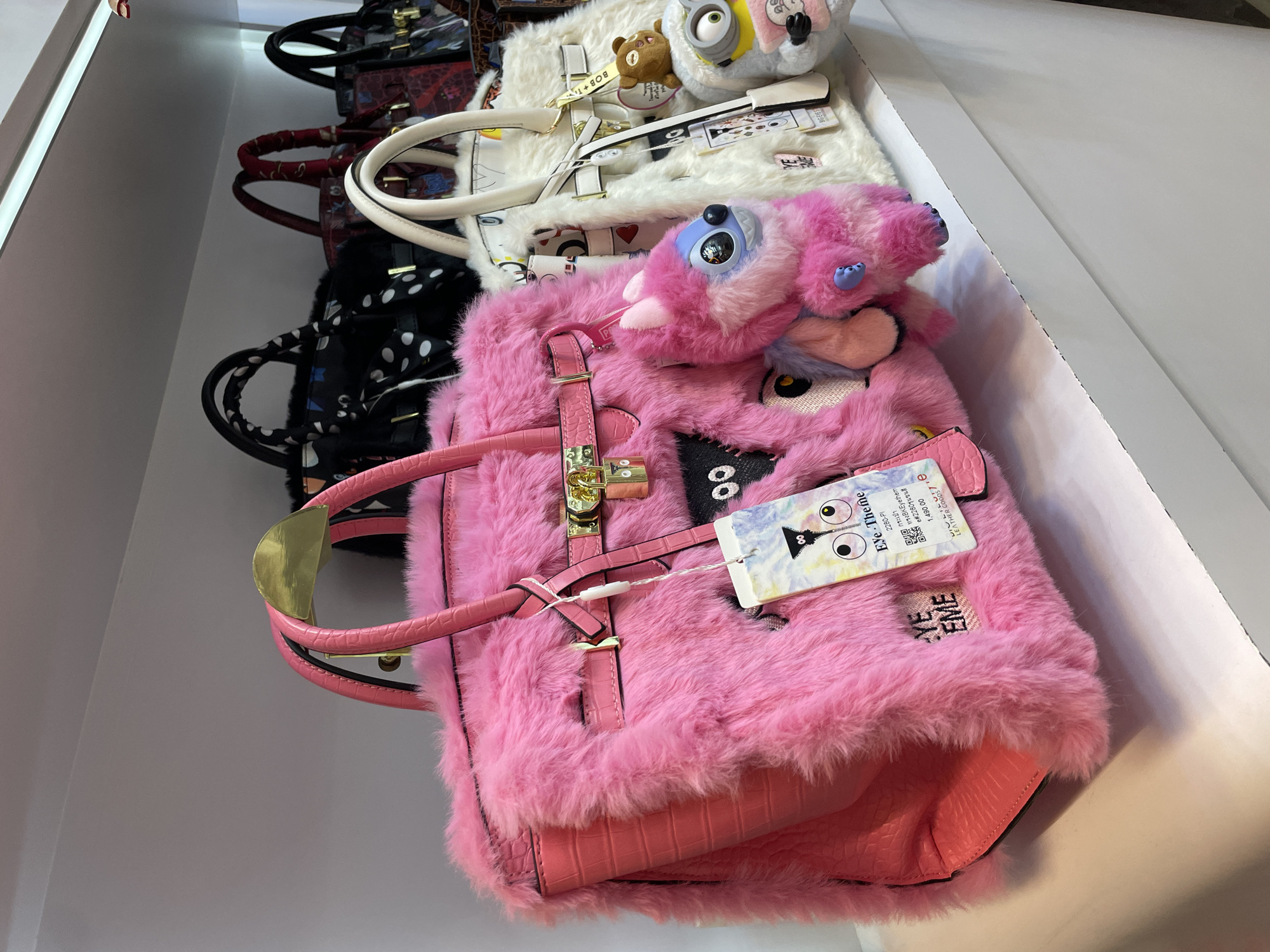 A close-up, eye-level shot shows a display of handbags on a white shelf. The foreground is dominated by a bright pink faux-fur handbag with a crocodile-textured pink leather base and handles. The bag is adorned with several patches, including a black square with white eyes, and a smaller black and white patch with a white eyeball. A plush toy, resembling a pink and blue creature with large blue eyes and purple paws, is attached to the handbag's strap. A white tag with a colorful design and text, including "Eye-Theme" and "Leather Goods," hangs from the bag.

Behind the pink bag, a white handbag with similar embellishments is visible. This bag also features patches, a gold lock, and a hanging tag. A small brown bear plush toy and a yellow Minion plush toy are attached to the white bag.

Further in the background, several other handbags are displayed. One is black with white polka dots, another has a red pattern, and a third appears to be dark red with a black pattern. The handbags are arranged neatly on the shelf. The lighting suggests an indoor retail environment. The scene is static and shows no people. This photograph was taken in Pattaya City, Thailand.