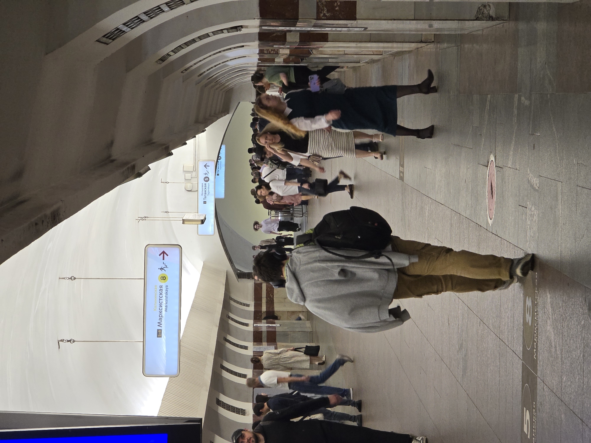 The image captures a bustling scene within a Moscow metro station. The station features a high, arched ceiling, a light-colored ceiling, and a tiled floor. Numerous people are walking in both directions along the platform. A large sign hangs from the ceiling, indicating "Марксистская/Marksistskaya" and directions to lines 5-6 and 8. Another smaller sign indicates Taganskaya lines 5 and 7. A man with a backpack faces away from the camera in the foreground.
