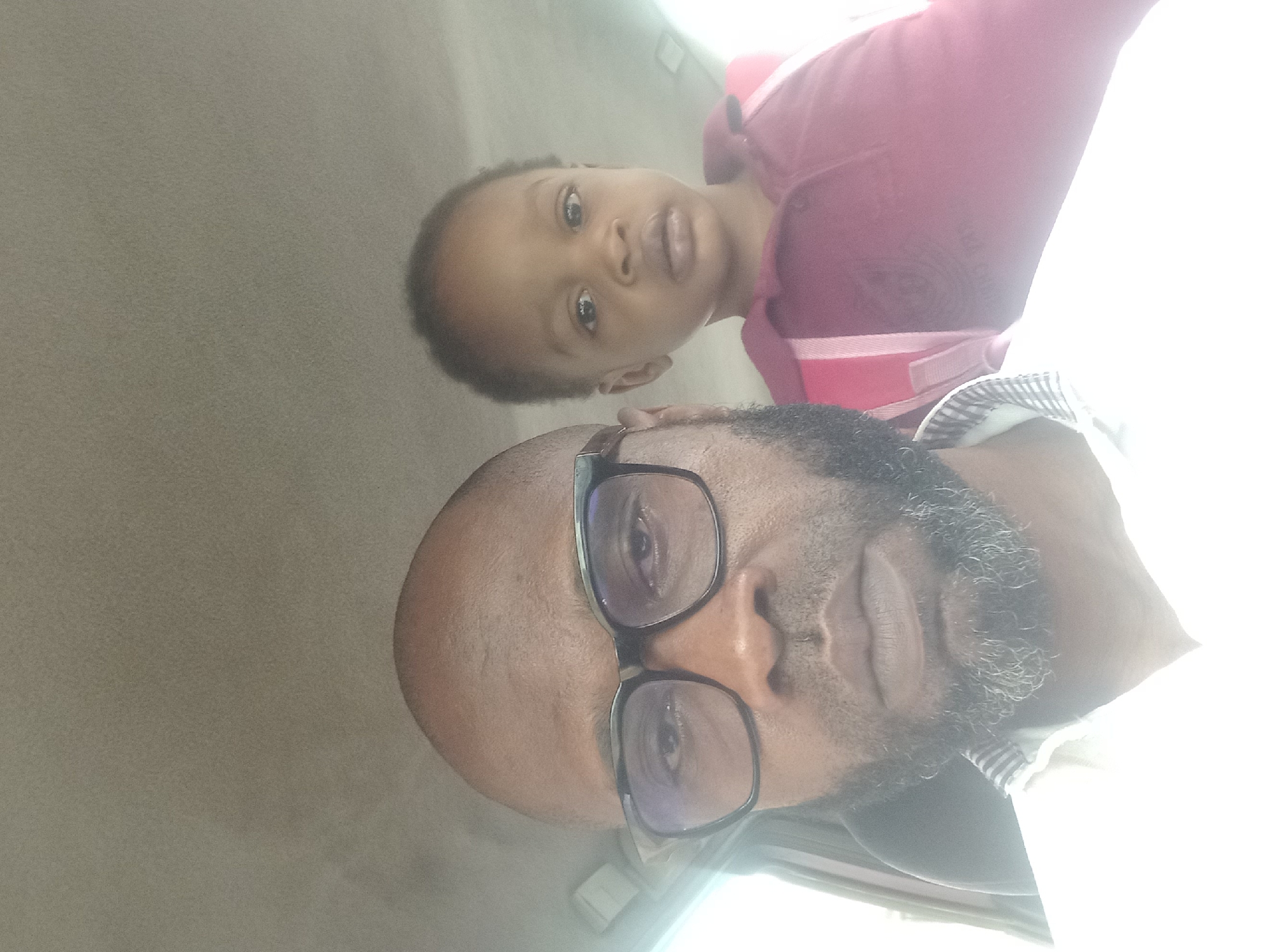 The image features an adult male and a young child, likely a father and child, captured in an indoor setting in Barden Yana, Nigeria. The photograph is taken from a slightly low angle, with the adult male in the foreground, and the child positioned behind and slightly above him. The adult male has a bald head and a full, dark beard with some visible grey strands. He wears dark-rimmed rectangular glasses, through which his eyes are visible, and appears to be wearing a collared shirt with a striped pattern on the cuff or collar area.
The child, appearing to be a toddler or young pre-schooler, has short dark hair and a light brown complexion. They are wearing a red or maroon polo shirt with some light-colored striping visible on the side. The child looks directly at the camera with a neutral, slightly curious expression and wide eyes. Both subjects have neutral emotions.
The background is a uniform, plain light gray or beige color, suggesting a simple wall or interior of a vehicle. A strong light source, possibly sunlight, enters from the right side of the frame, causing significant overexposure and glare, particularly on the right side of the adult's face, shirt, and parts of the background, indicating it is likely daytime. No text is visible in the image.