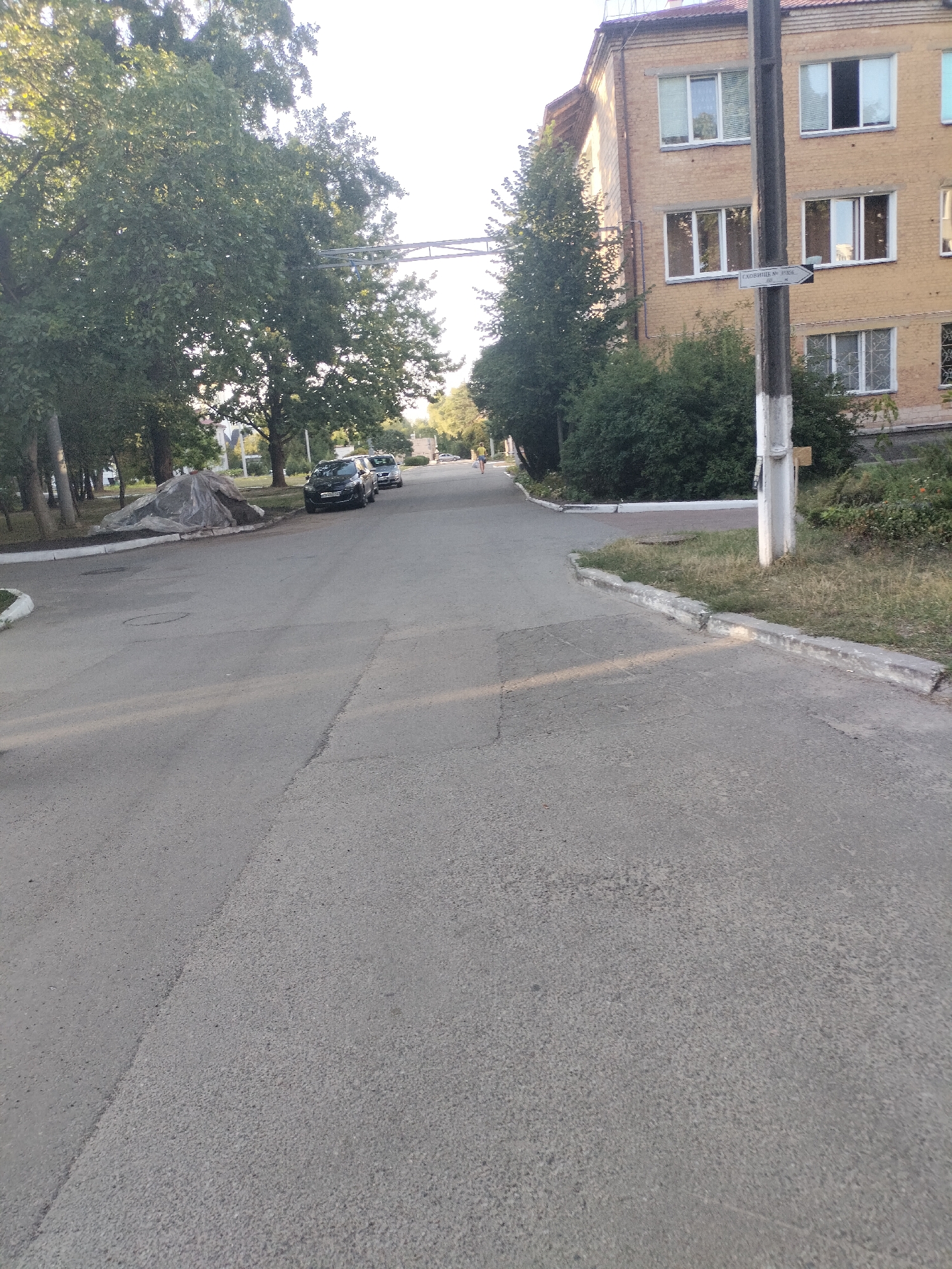 The image shows a street scene in Chernihiv, Ukraine. On the right side of the image is a multi-story brick building with several windows. A utility pole stands in front of the building, with a directional sign attached to it that reads "Сховище № 1". To the left of the building, there are trees and bushes. The street itself is paved with asphalt and has white painted curbs. Several cars are parked on the street. In the background, more trees and buildings are visible, and the sky is bright and overcast. A person is walking away from the camera on the sidewalk on the left side of the street.