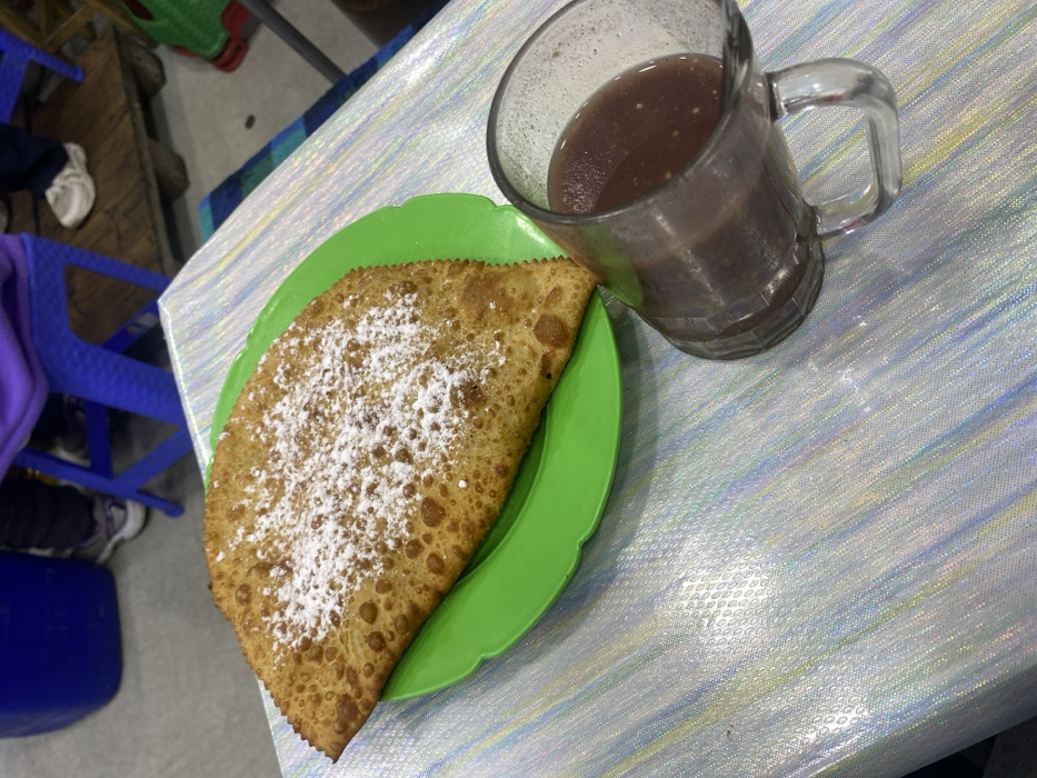 A fried turnover dusted with powdered sugar rests on a bright green plate, alongside a large glass mug filled with a dark, murky beverage. Both sit on a table with a light-colored, textured surface. Hints of chairs, including a bright blue one, are visible in the background. The composition is simple, showcasing the food and drink as the primary subjects.
