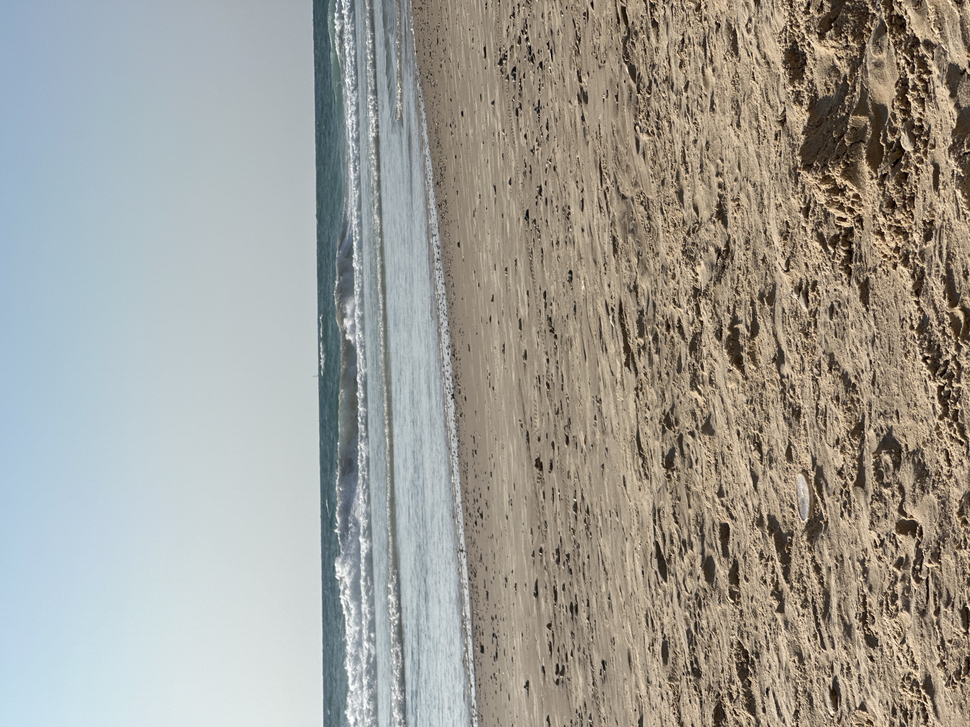 A wide shot of a sandy beach in Herzliya, Israel, shows the ocean meeting the shore under a clear sky. The foreground is dominated by textured, light brown sand with scattered small pebbles and indentations from waves or footsteps. The sand continues towards the water's edge, where a line of darker, wet sand marks the high tide line. Beyond this, the sea is a deep blue-green, with multiple rows of white-capped waves rolling towards the beach. The horizon is a sharp line separating the sea from the pale blue sky, which takes up the upper third of the image. There are no people or structures visible in the frame.