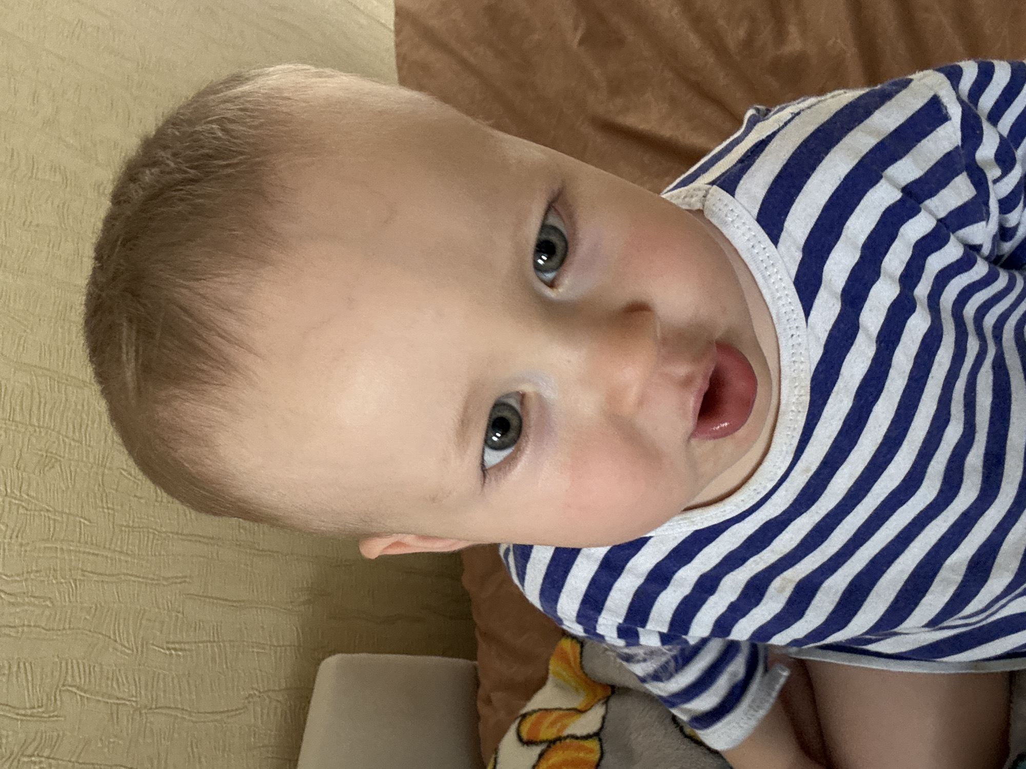 The image shows a close-up, overhead view of a young Caucasian child wearing a blue and white striped t-shirt. The child has light-colored hair and appears to be looking downwards with their mouth slightly open, revealing their tongue. A portion of a beige-colored, textured wall and a brown surface are visible in the background. To the left of the child, a gray plush toy with orange and white accents is partially visible. The scene is indoors. The location in the city of Naberezhnye Chelny, Russia cannot be determined from the visual information provided.