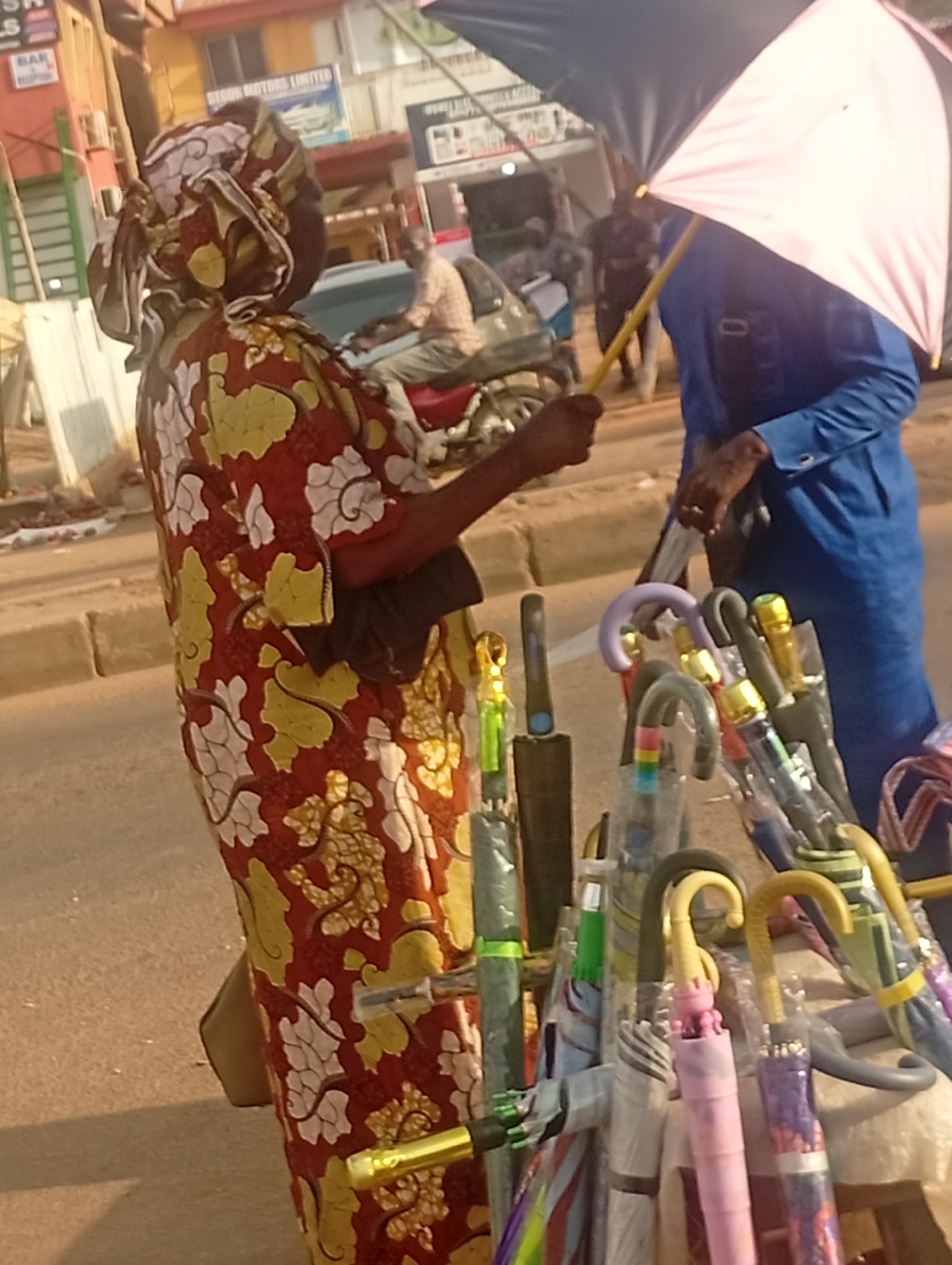 In Sabon Tasha, Nigeria, an umbrella vendor wearing a patterned red, gold, and white dress and head wrap is standing on the side of a street beside a display of umbrellas. A customer, dressed in blue and holding a pink-shaded umbrella, is present. Other individuals and vehicles are visible in the background.