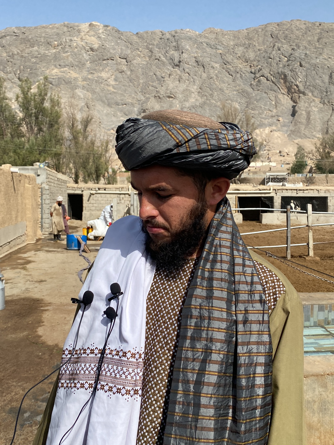 A man with a dark beard and turban stands outdoors. He is wearing a brown-patterned shirt, a shawl with gray and tan stripes, and a white embroidered cloth is draped over his shoulder, bearing three lapel microphones. In the background is a mountainous terrain, and what appears to be a small, rural structure. A man is visible in the background wearing a white robe and a blue container. The scene is in a rural, possibly agricultural setting. There is no clear evidence to confirm this photo was taken in Kandahar, Afghanistan.
