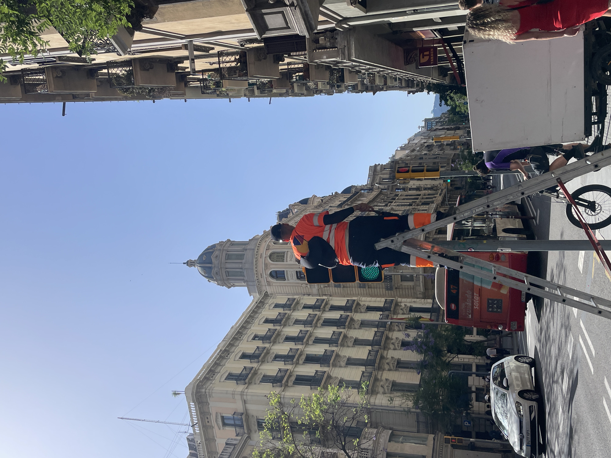 The image captures a street scene in Barcelona, Spain, under a clear blue sky, suggesting daytime with fair weather. In the foreground, a man wearing an orange high-visibility vest and dark pants is working on a ladder, positioned in front of a traffic light. The traffic light is green, indicating that traffic can proceed. The ladder is extended upwards, and the man appears to be engaged in some form of maintenance or repair work.
Behind the man and the ladder, several buildings with ornate architecture line the street. One building features a distinctive dome, characteristic of some European city designs. There's a red bus with the number "47" visible on its rear and text that appears to read "ciudad solar." A white car is parked on the street. Further in the background, more buildings with balconies and windows are visible, giving a sense of an urban environment.
In the upper right of the image, part of a white structure, possibly a truck or a large vehicle, is visible, with a person in a red top and another in a purple and black outfit standing near it. A bicycle is also present next to the ladder. The overall impression is a busy urban street with ongoing work, likely related to traffic control or infrastructure. The sunlight suggests it's a bright, possibly warm, day.