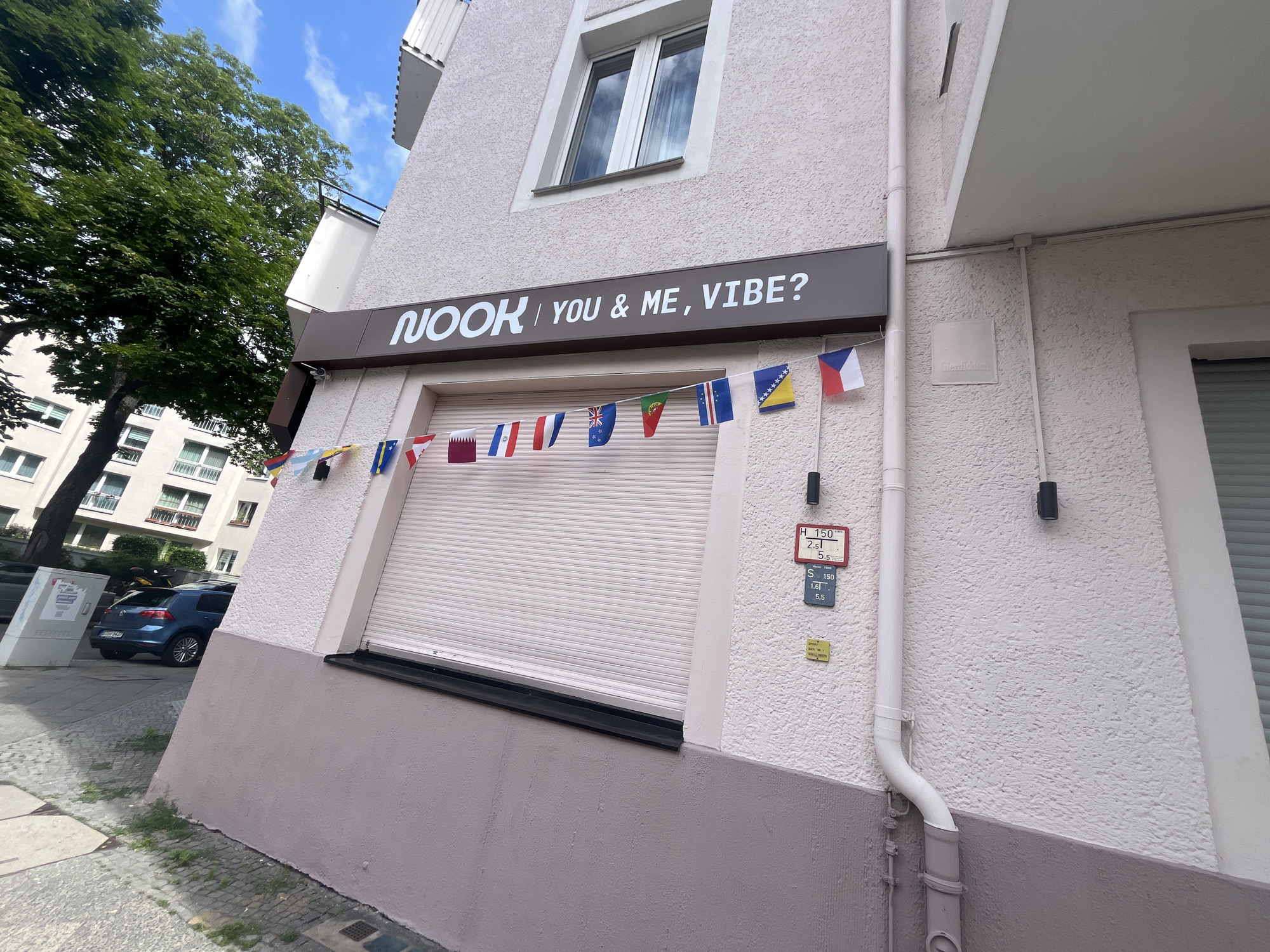 A low-angle, eye-level outdoor shot captures the exterior of a commercial building in Berlin, Germany, on a bright, cloudy day. The building's facade is textured and painted in a pale pinkish-gray hue, with a darker pink base. A white downspout runs vertically along the wall. Above the downspout, there are two red and blue rectangular signs with white text, possibly indicating building or utility information. A string of small, colorful international flags hangs across the front of the building above a closed, light-colored rolling security shutter. Above the shutter, a dark brown sign with white lettering reads "NOOK | YOU & ME, VIBE?". A window with a white frame is visible on the upper floor. To the left of the building, a large green tree with dense foliage stands tall, and part of a multi-story residential building with balconies and windows is visible behind it. On the street in front of the commercial building, a blue hatchback car is parked. The sidewalk is paved with cobblestones and concrete slabs, with patches of green grass growing between them.