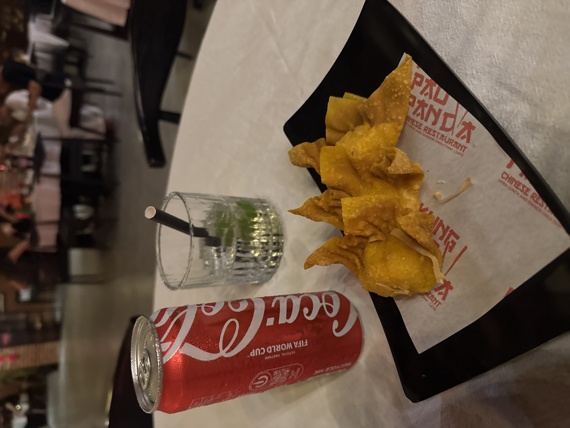 The image shows a table at a Chinese restaurant, likely in Kuta, Indonesia, with a meal consisting of fried wontons, a can of Coca-Cola, and a glass of water with a straw. The wontons are golden brown and arranged on a black rectangular plate lined with paper that has red Chinese characters and English text that reads "PANDA CHINESE RESTAURANT" and "OPEN LUNCH AND DINNER DAILY 11AM - LATE". The Coca-Cola can is red with the iconic white script logo and the text "FIFA WORLD CUP™ OFFICIAL PARTNER". The can is covered in condensation, suggesting it is cold, and the glass of water also has condensation and contains ice and what appears to be lime slices, with a black straw. The table is white, and the background is blurred, showing what appears to be other diners and restaurant furniture, indicating an indoor setting during daylight or under artificial light. The scene evokes a casual dining experience, possibly a lunch or dinner outing.