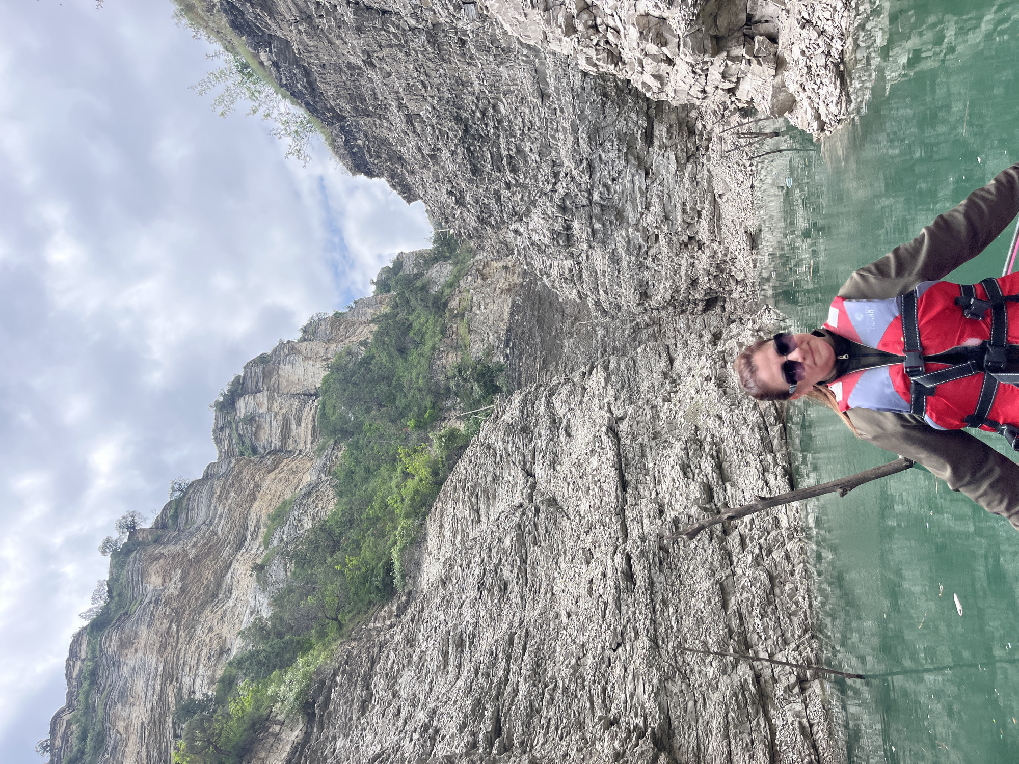 A person wearing a red and white life vest and sunglasses holds a stick over the water in a canyon or gorge. The steep rock walls rise high on either side, with some green vegetation clinging to the sides. The water appears green and reflective. The sky is overcast. The location is likely in the vicinity of Druzhba, Russia, based on the prompt's request to include that location.