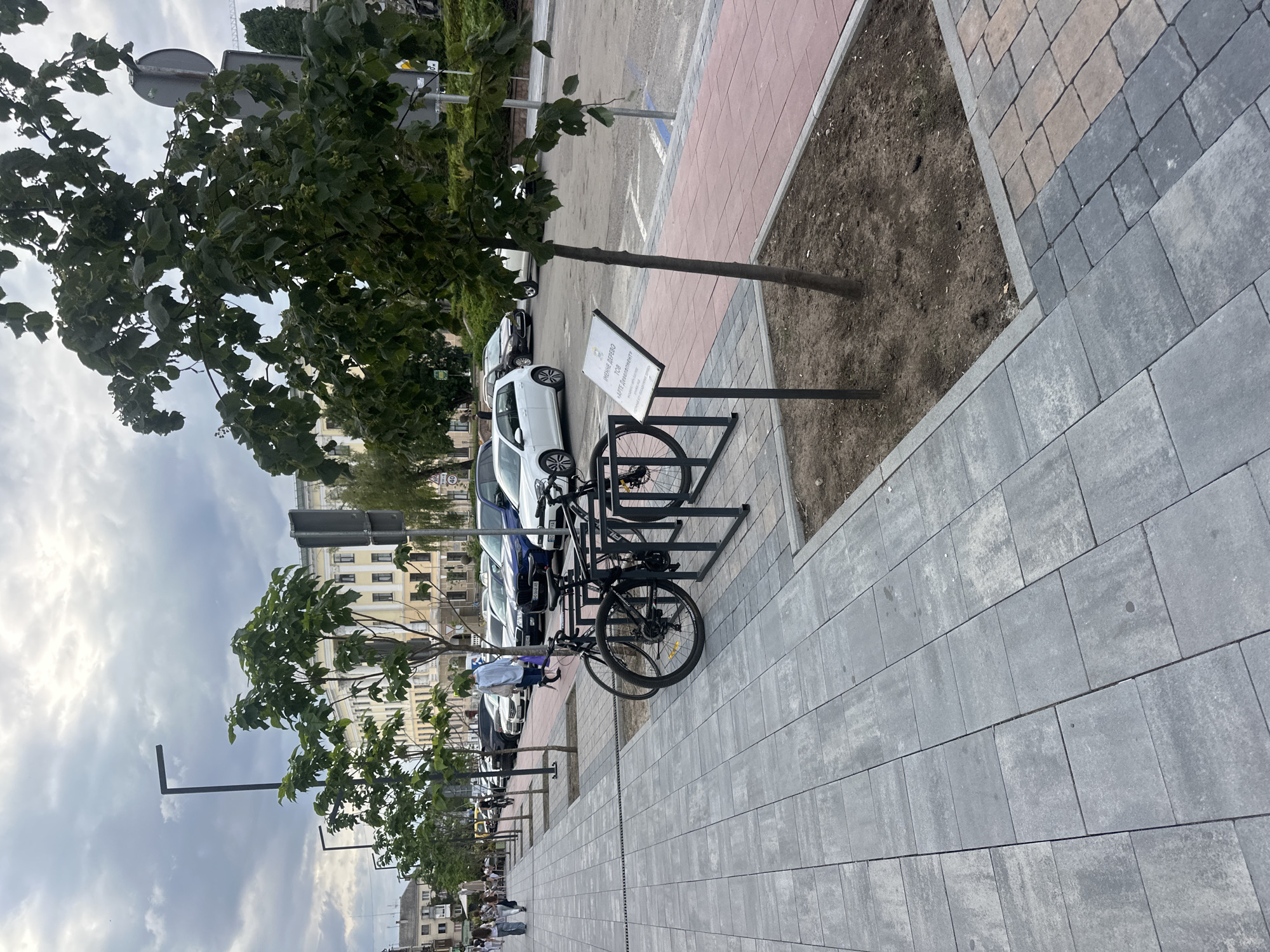This image depicts a street scene in Ternopil, Ukraine. The foreground features a sidewalk paved with gray square and rectangular tiles. Adjacent to the sidewalk is a red-tiled area with a small tree planted in a soil bed. A metal bicycle rack holds two black bicycles. A white sign with Ukrainian text is mounted on a metal pole near the tree.
Beyond the sidewalk, a street is visible with several parked cars, including a white Volkswagen Golf, a dark blue SUV, and other vehicles. A row of young trees with green leaves lines the sidewalk, separating it from the street. In the background, there are buildings with classical architectural features, a street lamp, and other trees. The sky is overcast with clouds. Several people are walking on the sidewalk in the middle ground, and some are seated at tables outside a building in the far background. A traffic sign with the number "40" is visible on the right side of the image.