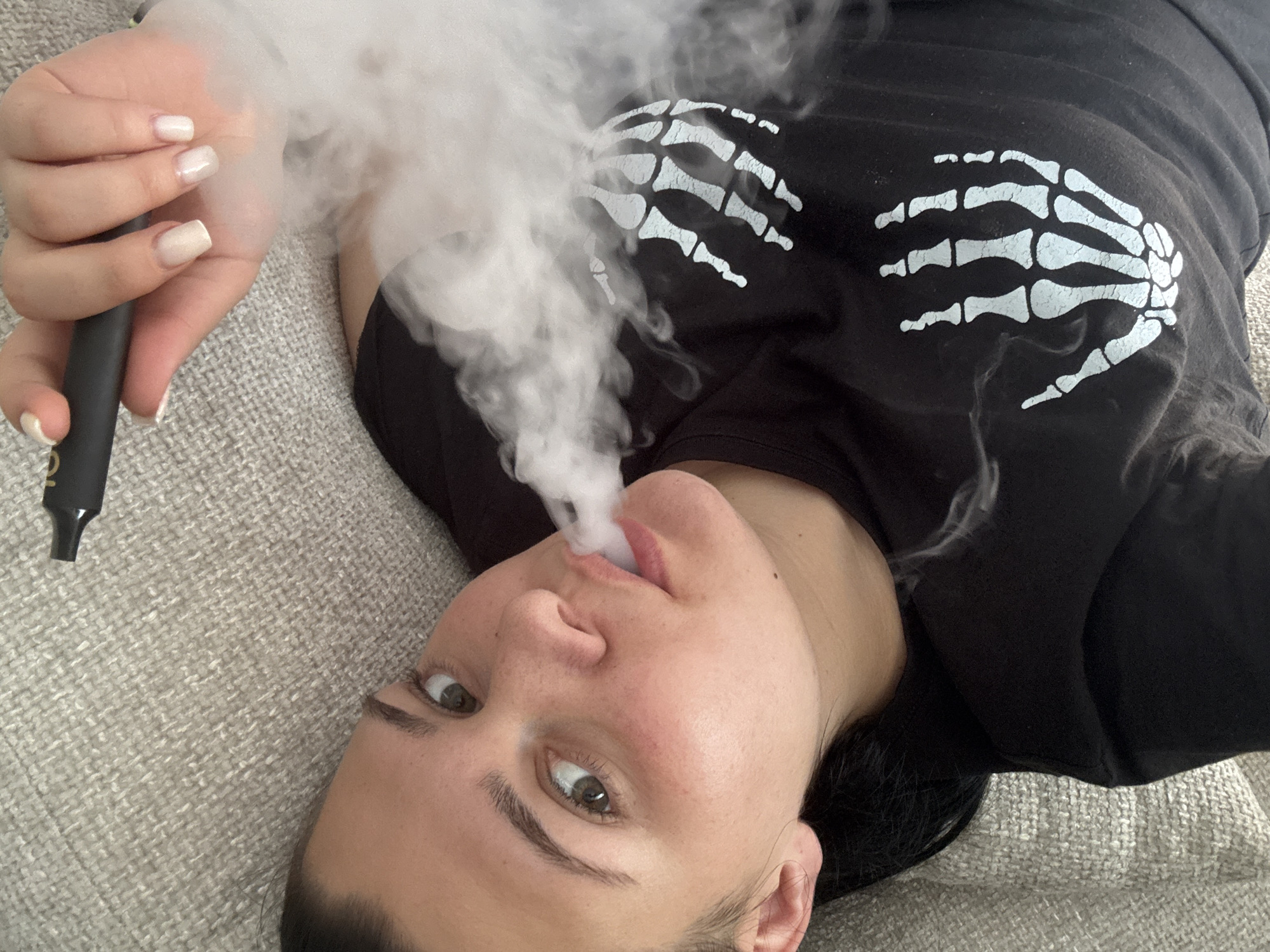 The image presents a close-up, diagonal view of a person lying down indoors in Rogosh, Bulgaria. The individual, whose face is angled upwards towards the camera, is exhaling a large, dense cloud of white vapor from their mouth, partially obscuring their chin and the area around their right hand. They have light brown eyes and dark eyebrows, maintaining a neutral expression while looking directly at the viewer.
Their right hand, featuring light-colored, neatly manicured square-tipped nails, holds a sleek black, pen-shaped vaping device. A faint, possibly gold-colored logo, appearing to read "VO" or "YO," is visible on the side of the device. The person is wearing a black long-sleeved shirt adorned with a prominent white graphic of a skeleton hand on the upper arm/shoulder area. They are resting on a light grey, textured fabric surface, indicative of a sofa or bed in an indoor setting. The image captures the moment of exhaling vapor from the device.