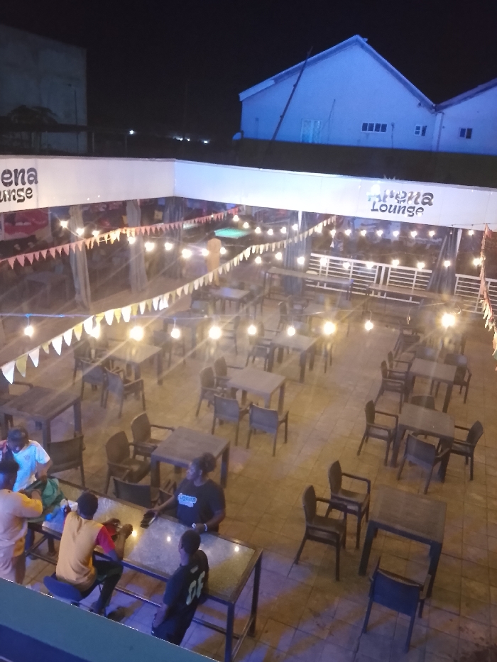 The image displays an outdoor lounge area identified by signage as "Athena Lounge," located in Narayi, Nigeria, at night. The scene is illuminated by numerous overhead string lights featuring exposed bulbs and triangular bunting.
The primary area consists of multiple dark-colored square and rectangular tables, predominantly unoccupied, each accompanied by several matching chairs, arranged on a light-colored paved surface.
Six individuals are visible. In the lower-left foreground, four persons are clustered around a rectangular table with a glass top. One individual in a light-colored shirt is standing, leaning over the table. Another, in an orange short-sleeved top, stands adjacent. A third, wearing a yellow shirt, is seated on a dark stool, engaged with a mobile device. A fourth, wearing a dark t-shirt with "CF" on the back, is also seated at this table. Towards the center-left, a person with dark, curly hair, wearing a black t-shirt with "Athena Lounge" printed on it, stands near an empty table, holding a phone. A sixth individual is partially visible standing near a green-felt pool table in the upper-left midground.
In the background, under a dark night sky, prominent white buildings with pitched roofs are visible. White railings demarcate elevated sections of the lounge.