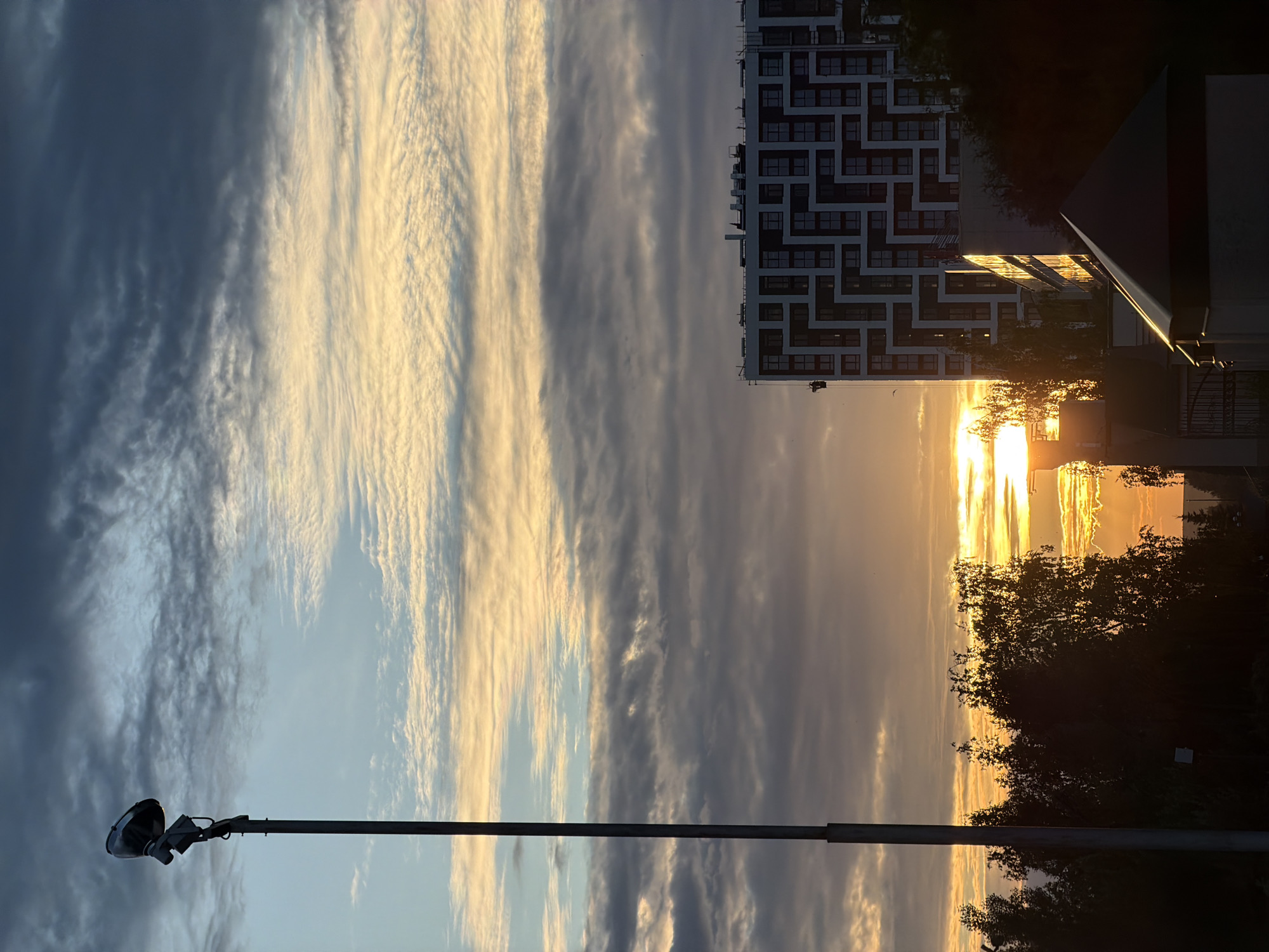 The image depicts a dramatic sunset over an urban landscape, likely in Nizhnekamsk, Russia. The sky is filled with a striking contrast of dark, streaky clouds on the left and bright, golden clouds illuminated by the setting sun on the right. The sun itself is a brilliant orb peeking through the horizon, casting a warm glow that reflects off a modern, multi-story building with a distinctive geometric facade.
In the foreground, a tall lamppost with a single, dark lamphead is positioned vertically, its stark silhouette cutting across the colorful sky. Trees, silhouetted against the bright sunset, frame the right side of the scene, adding a natural element to the urban setting. The building appears to be residential or commercial, with numerous windows visible.
The time of day is clearly sunset, indicated by the low angle of the sun and the vibrant, warm colors in the sky. The weather appears to be clear with scattered clouds, creating a visually dynamic atmosphere. The overall mood is one of awe and beauty, with the contrasting elements of the natural sky and the man-made structures creating a striking composition. No visible text is present in the image.