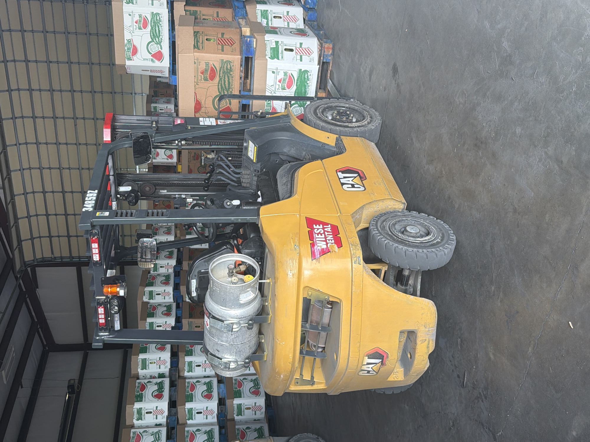 A yellow Caterpillar forklift is parked indoors in a warehouse setting in Hornersville, United States. The forklift is positioned in front of stacks of cardboard boxes filled with watermelons, some of which are labeled "Fresh Watermelons." The warehouse has a concrete floor and a large overhead garage door in the background. The forklift is equipped with a propane tank mounted on the back. The overall environment suggests a distribution or storage facility for produce. There are no people visible in the image.