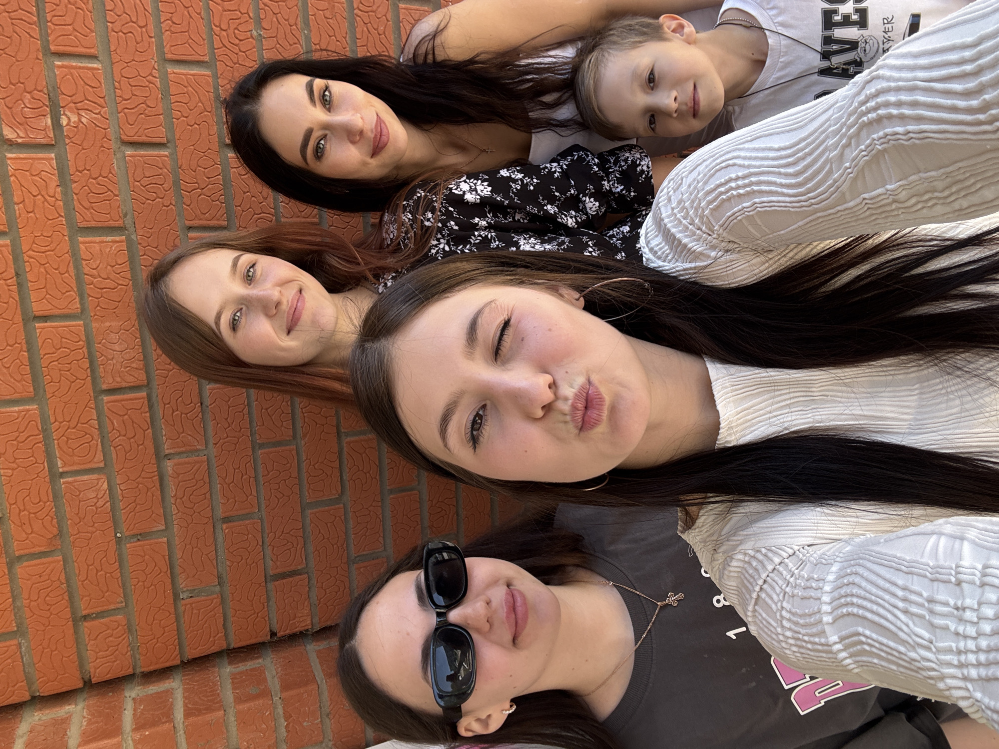A group of five individuals are posing for a selfie against a brick wall, presumably outdoors, with the wall taking up the left side of the frame. Four are females and one is a male child. Starting from the bottom, a woman with sunglasses and a gray shirt is looking at the camera. Above her is a woman winking and making a kissing face while wearing a white top. Above these two, another woman with brown hair is smiling. Adjacent to her, towards the top, a woman with dark hair is looking at the camera. Next to her is a child wearing a white shirt with a graphic. Based on the information provided, this photo was taken in Chaikovsky, Russia.