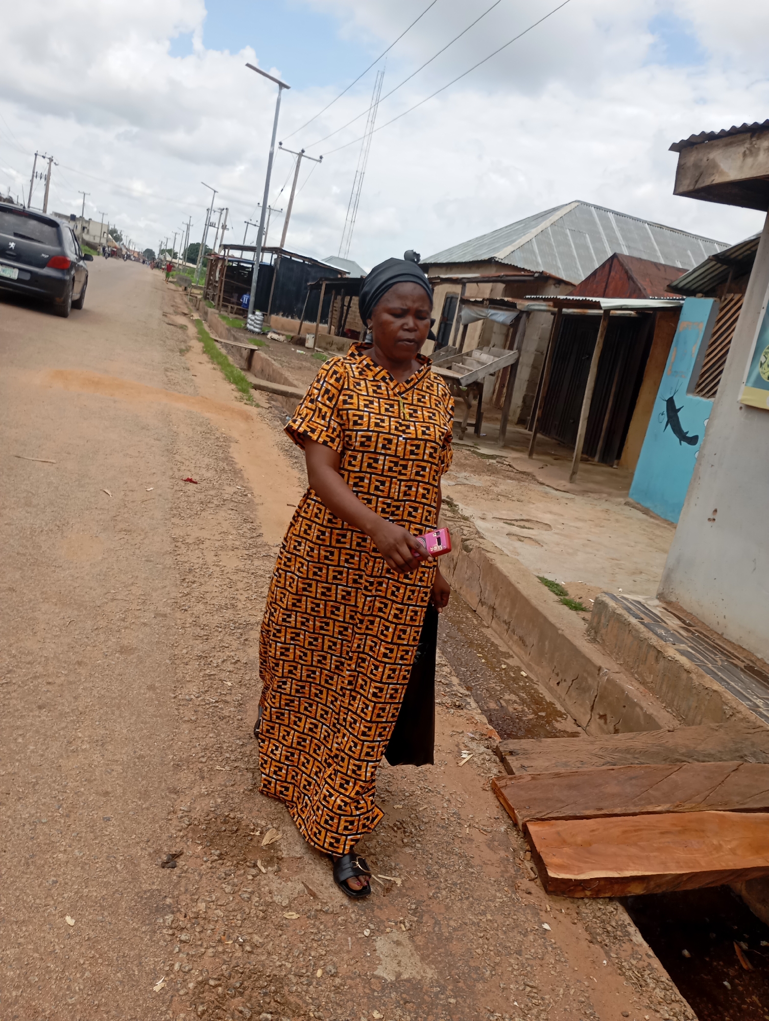 In Shendam, Nigeria, a woman wearing a black headwrap and an orange and black patterned dress walks on a dirt road. She holds a pink mobile phone in her right hand and a black bag in her left. She is wearing black sandals. The road is lined with a concrete drainage ditch and various structures, including corrugated metal-roofed buildings and stalls. A black car is parked on the left side of the road. The sky is cloudy.