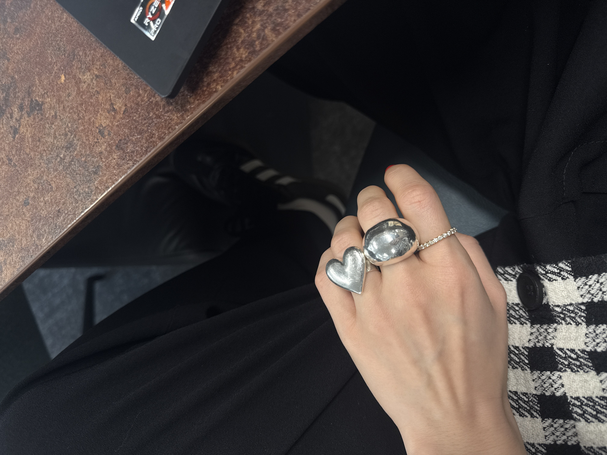 A high-angle shot shows a woman's hand with silver rings resting on her lap. The rings are on the middle and ring fingers; one ring is shaped like a heart, and the other is a rounded dome. A third ring with small stones is on the pinky finger. The woman wears black pants and a black and white checkered jacket. A desk with a dark wood-grained surface is visible in the upper left corner, with a laptop with an AMD Ryzen Pro logo on it. The background shows a shoe with Adidas stripes on the floor. The location appears to be indoors and is not recognizably in London.