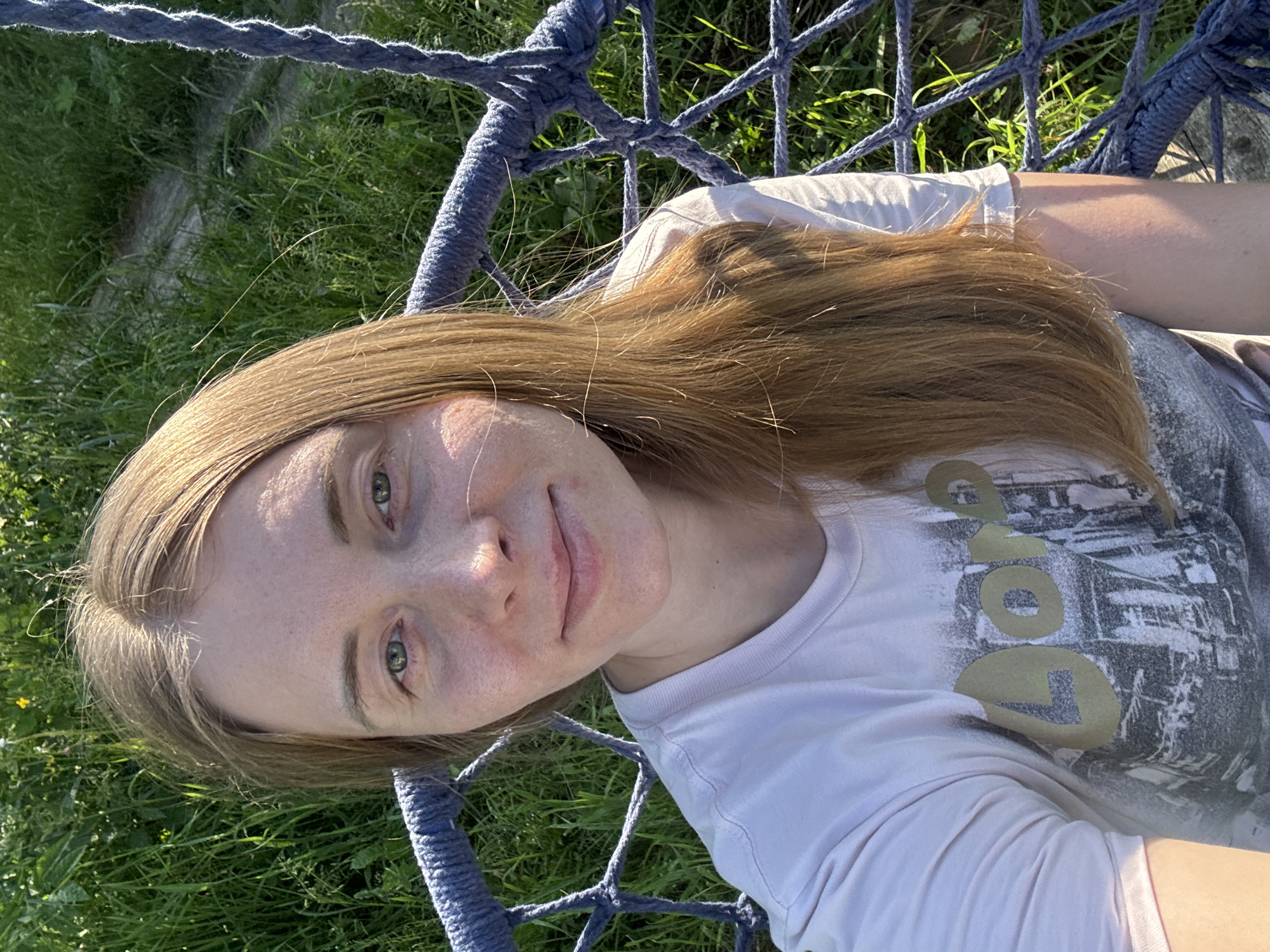 A young woman with long, auburn hair is sitting in a blue rope swing outdoors, with a background of green grass and vegetation. She is wearing a white t-shirt with a graphic print. Her head is tilted slightly, and she is looking towards the camera with a subtle smile. The lighting suggests it is daytime, with sunlight casting shadows on her face and clothing. The scene appears to be in a natural outdoor setting, possibly a park or a backyard. There is no indication of the city of Kurgan, Russia.