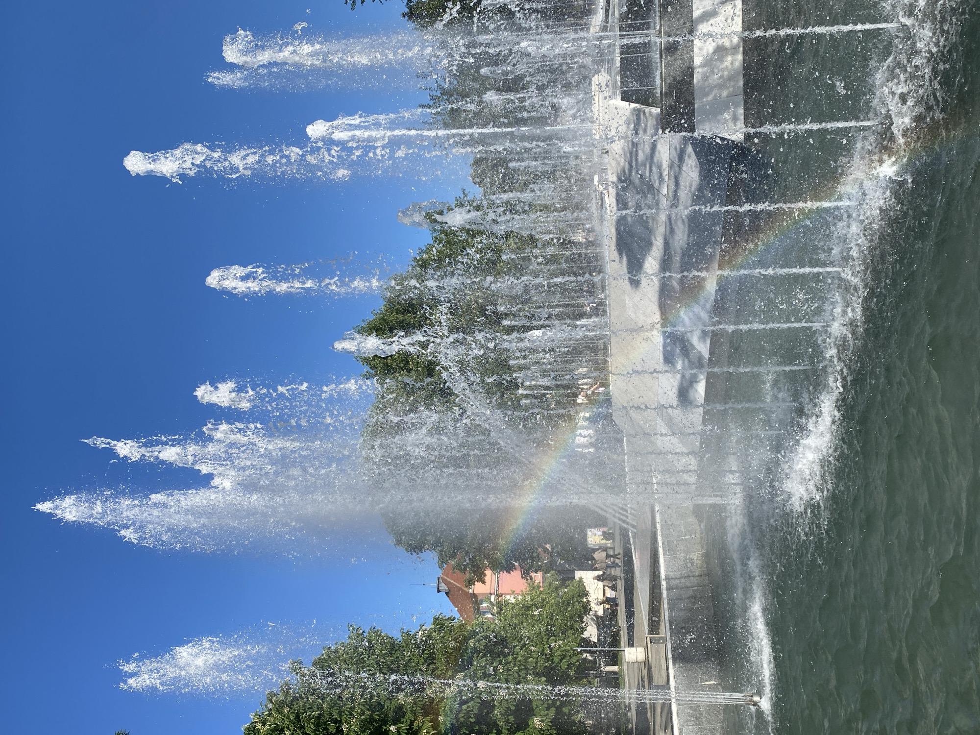 The image captures a fountain in Pleven, Bulgaria. The fountain features multiple jets of water shooting upwards against a clear blue sky, with trees visible in the background. A rainbow is visible in the mist of the fountain, creating a colorful arc. The fountain's architecture includes stone or concrete structures, and there's water pooling below. People can be seen walking along the edge of the fountain. A building with a reddish-orange roof is seen in the background.