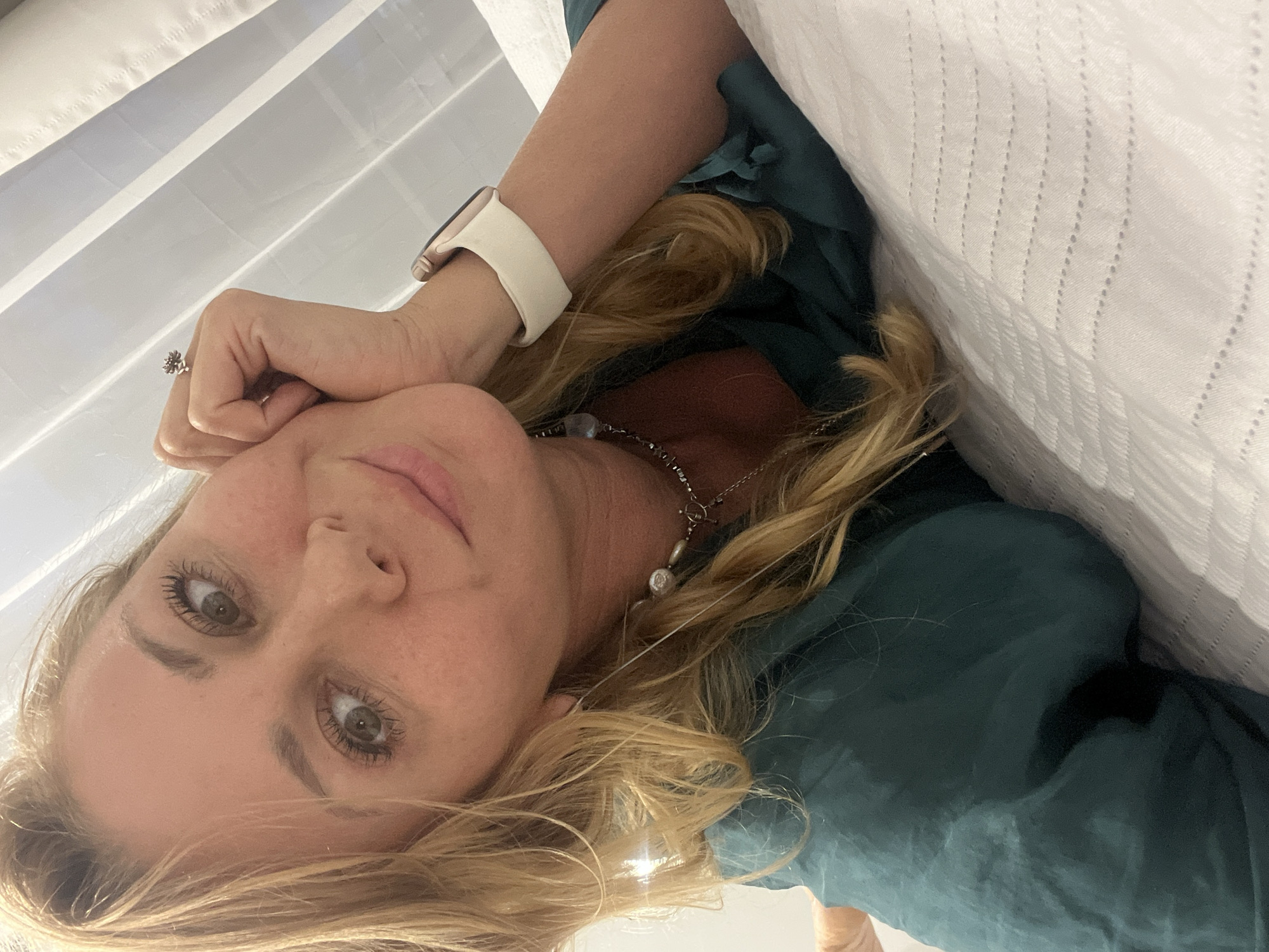 A woman with blonde hair and fair skin is looking at the camera. She is wearing a dark green top and a white watch. She has on a silver necklace. Her head is resting on her hand, which has a ring on the ring finger. A white textured fabric is in the background. The location cannot be determined.