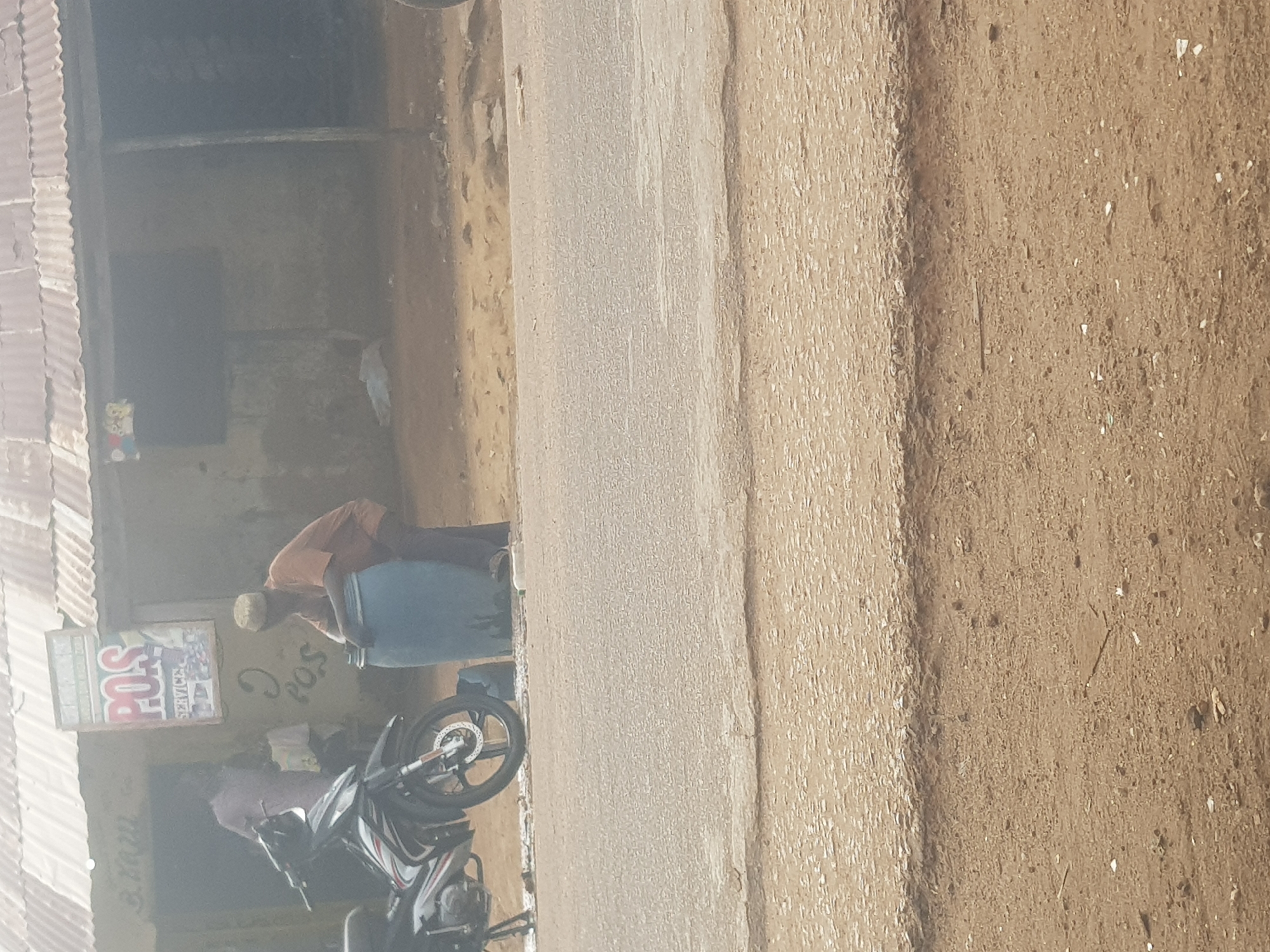 The image displays an outdoor street scene in a semi-urban or rural setting, likely Kufena, Nigeria, under bright daytime conditions. A paved road runs horizontally across the middle ground, bordered on the right by a wide, light brown dirt or sand shoulder. On the left side of the road, there is a building with a corrugated metal roof and light brown, textured walls.
In front of this building, a man wearing an orange or brown shirt and a white skullcap (kufi) is bent over a large blue plastic barrel, appearing to be engaged in an activity like filling or dispensing. Below the barrel and slightly to its left, a black and white motorcycle with visible disc brakes is parked. Attached to the building's wall, above the man, is a rectangular sign prominently displaying "POS SERVICE." Below this sign, the word "POS" is also scrawled directly onto the wall. A second person is partially obscured behind the parked motorcycle.
The scene depicts a typical roadside environment, possibly a local shop or service point, with bright, even lighting consistent with a sunny day. No strong shadows are cast, indicating either high sun or diffuse light.