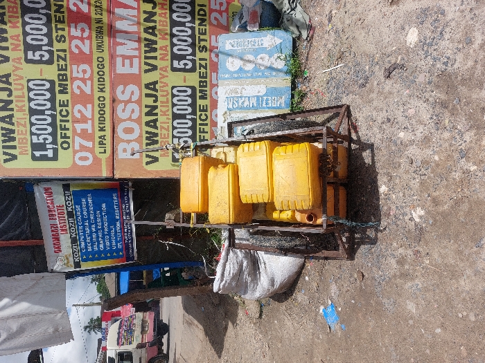 The image depicts an outdoor scene in Dar es-Salaam, Tanzania, specifically referencing Mbezi and Kiluvya areas based on prominent signage. In the foreground, a two-wheeled, unpainted metal handcart is positioned on dry, uneven dirt and gravel ground. The handcart carries a stack of at least five rectangular yellow plastic jerrycans, some secured with blue cordage. To the left of the handcart, a large, partially filled white woven sack rests on the ground.
Dominating the mid-ground are several large advertising banners. On the left, a white banner with blue text advertises "OKOMBOZI EDUCATION INSTITUTE," listing courses such as "COMPUTER COURSE," "SECRETARIAL COURSE," "GRAPHIC DESIGN," and "VIDEO PRODUCTION." To its right, two large yellow and green banners, stacked vertically, advertise "VIVANJA" (plots of land) in "MBEZI, KILUVYA NA KIBAHA," citing prices ranging from "1,500,000 - 5,000,000" and providing a contact number "0712 25 25 25." The lower banner specifically names "BOSS EMMA."
In the background, on the left, a white and pink minivan (locally known as a dala-dala) is visible on a paved road, facing away from the viewer. Other indistinct structures and some green foliage are present further in the background. No people are visible in the scene. The lighting suggests bright daylight, casting distinct shadows from the handcart and other objects onto the ground. Scattered debris, including a blue plastic sheet, is visible on the ground.