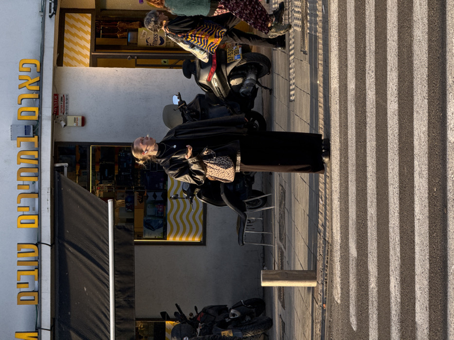 The scene depicts an urban street in Tel-Aviv, Israel, during late afternoon with long shadows cast by sunlight from the right. In the immediate foreground, a pedestrian crosswalk with white stripes spans an asphalt road. A light-colored cylindrical bollard is situated on the paved sidewalk to the left of the crosswalk.
The primary subject, a light-skinned adult female with blonde hair pulled back, stands in the midground on the sidewalk, facing slightly right of the camera. She wears a black turtleneck, a loose black leather jacket, a long black skirt extending to her ankles, and black shoes. She carries a leopard-print shoulder bag on her left shoulder and wears a silver pendant necklace and an earpiece in her right ear. Her right hand holds a small, light-colored object to her lips, while her left hand is open, palm up, at waist level.
Behind the primary subject, two other individuals are partially visible walking from right to left. The person closer to the camera wears a patterned orange, yellow, and black top, dark trousers, and carries a multicolored tote bag. The person further back wears a green top and a floral-patterned skirt.
Several scooters are parked along the sidewalk in the midground and background. One silver-colored scooter clearly displays an Israeli license plate: "97-530-30". A black chair with metal legs is positioned near one of the parked scooters.
The background features a white storefront facade. Above a dark retractable awning, a large yellow sign with Hebrew lettering reads "פארם ובתי מרקחת טובים" (Pharm and good pharmacies). An alarm box with Hebrew text, including "אזהרה" (Warning) and contact numbers, is mounted on the wall to the right of the awning. To the far right, a store entrance features a distinctive yellow-and-white zigzag pattern on the wall and a visible "Fitness" sign.