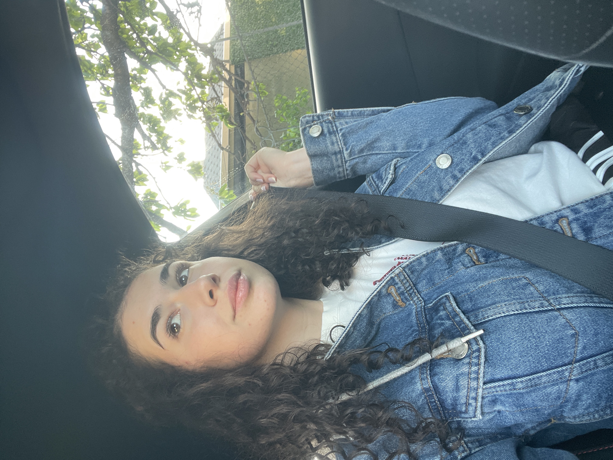 The image features a young woman with long, dark, curly hair, lying or reclining in what appears to be a car seat. She has a fair complexion, dark eyebrows, and is looking directly at the camera with a neutral and somewhat relaxed expression.
She is dressed in a blue denim jacket with silver buttons, worn over a white t-shirt. A dark car seatbelt crosses her chest. Her left hand is visible, showing neatly manicured nails with a French tip style.
The setting is inside a vehicle. Through the car window, bright daylight illuminates lush green tree branches, suggesting an outdoor environment. Faintly visible in the background through the window are hints of a fence or a building structure. The interior of the car, including the seat and roof lining, is dark-colored.
The time of day is clearly daylight, indicated by the bright sun illuminating the trees outside. The activity is a posed photograph, with the woman looking towards the lens. There are no clearly discernible words or text visible on her clothing or in the background. The overall impression is casual and serene.