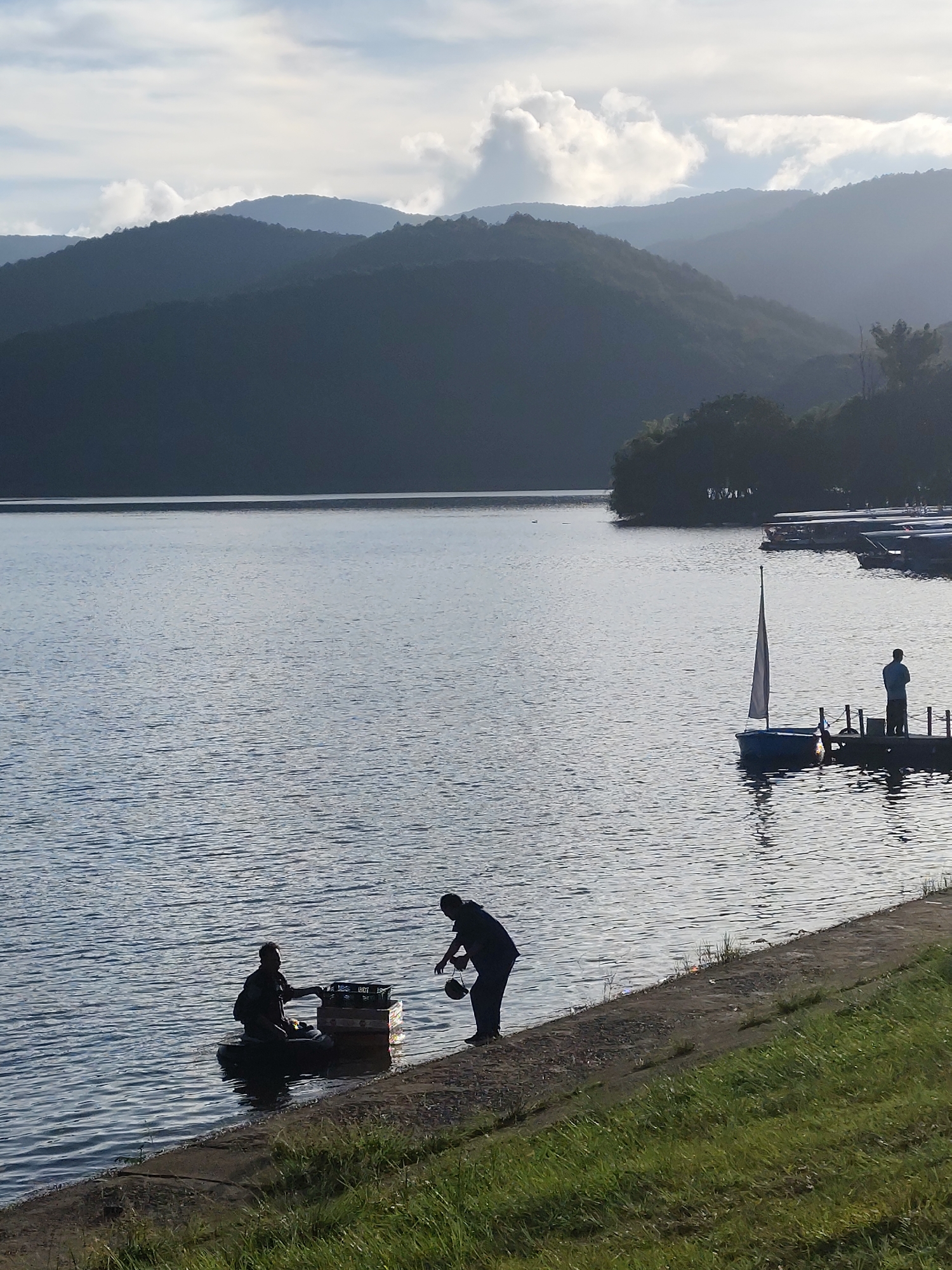 The image depicts a tranquil scene on Tuyen Lam Lake in Da Lat, Vietnam. In the foreground, two silhouetted figures are interacting by the water's edge. One person is on a makeshift raft or inner tube loaded with containers or boxes, while the other, standing on the bank, appears to be handing something to the person on the raft.
Further into the lake, on a small pier, stands another silhouetted figure. A small sailboat is tied to the pier. In the distance, across the lake, are forested hills. Moored boats line the far shoreline. The sky is overcast with clouds.