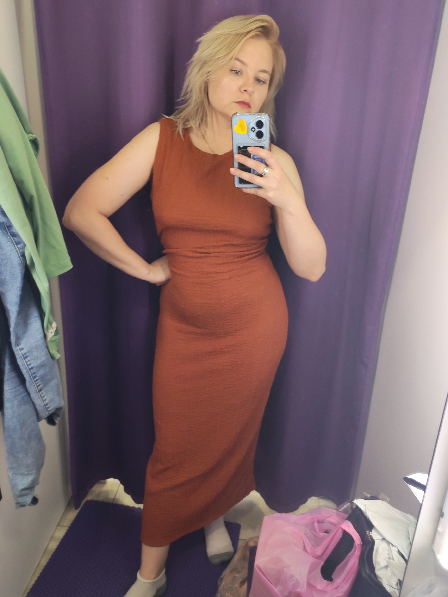 A woman is taking a selfie in a dressing room. She is wearing a rust-colored sleeveless dress that falls below the knee, and white socks. The dressing room has a purple curtain behind her and a purple floor mat. Clothing items are visible on the left and lower right of the frame. The woman is holding a blue phone with a yellow heart sticker on it. The location is a dressing room, but specific location in Izhevsk, Russia cannot be determined from the image.