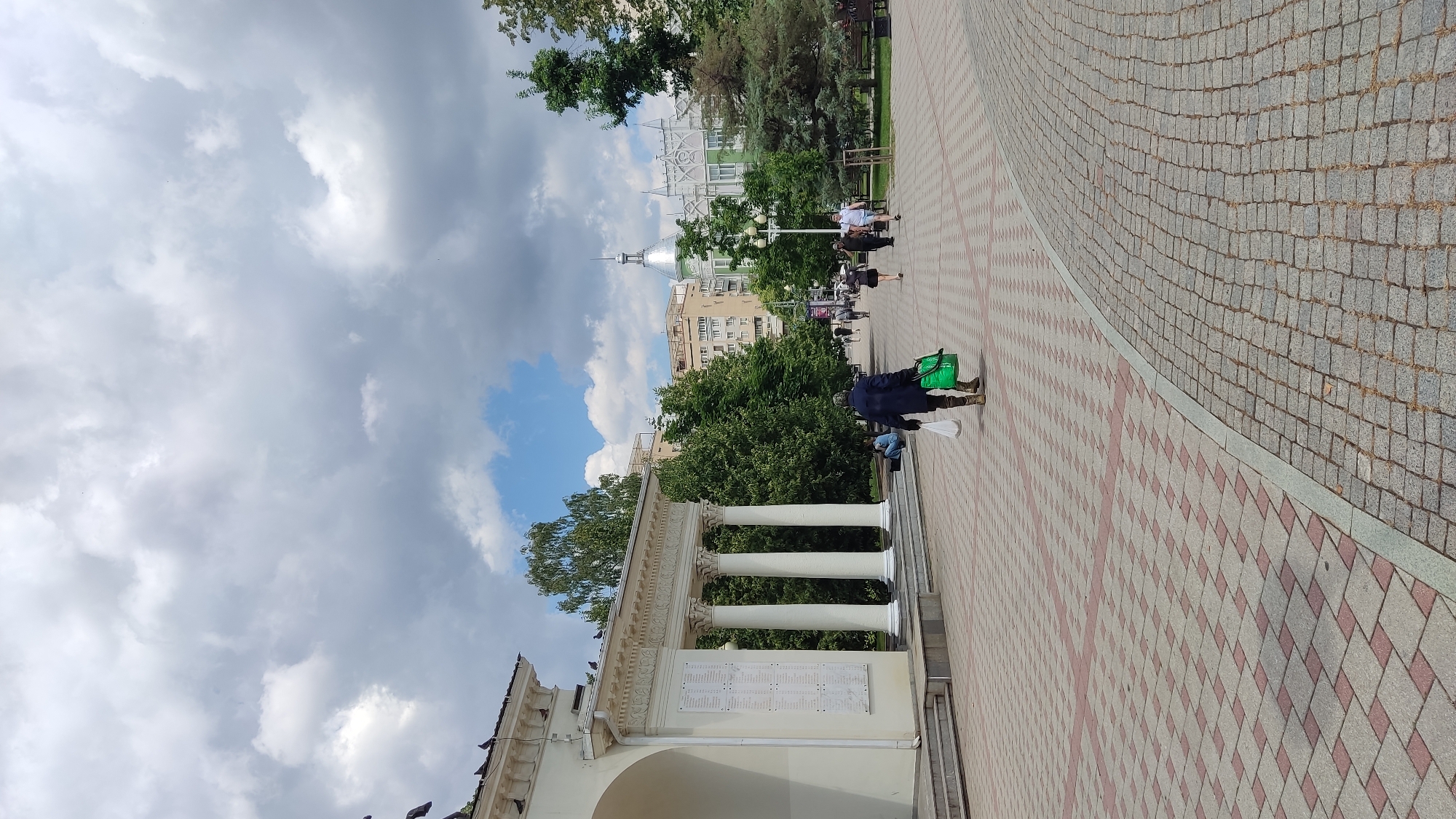 The image depicts a pedestrian walkway in a park-like urban setting, likely in Krasnodar, Russia, given the architectural styles and greenery. In the foreground, a person walks away from the camera, carrying a white plastic bag in their left hand and a green tote bag in their right. The person is wearing dark clothing and brown boots. The walkway is paved with a pattern of square red and grey tiles. To the left, a white building with a colonnade of four large columns is visible. Pigeons are perched on the roof of this building. In the background, various trees line the walkway, and several buildings of different architectural styles are visible, including a light-colored building with a green, domed roof and intricate white facade. Other people are scattered throughout the scene, some walking and others sitting. The sky is partly cloudy with dramatic grey and white clouds. The scene suggests a typical summer day in a public urban space.