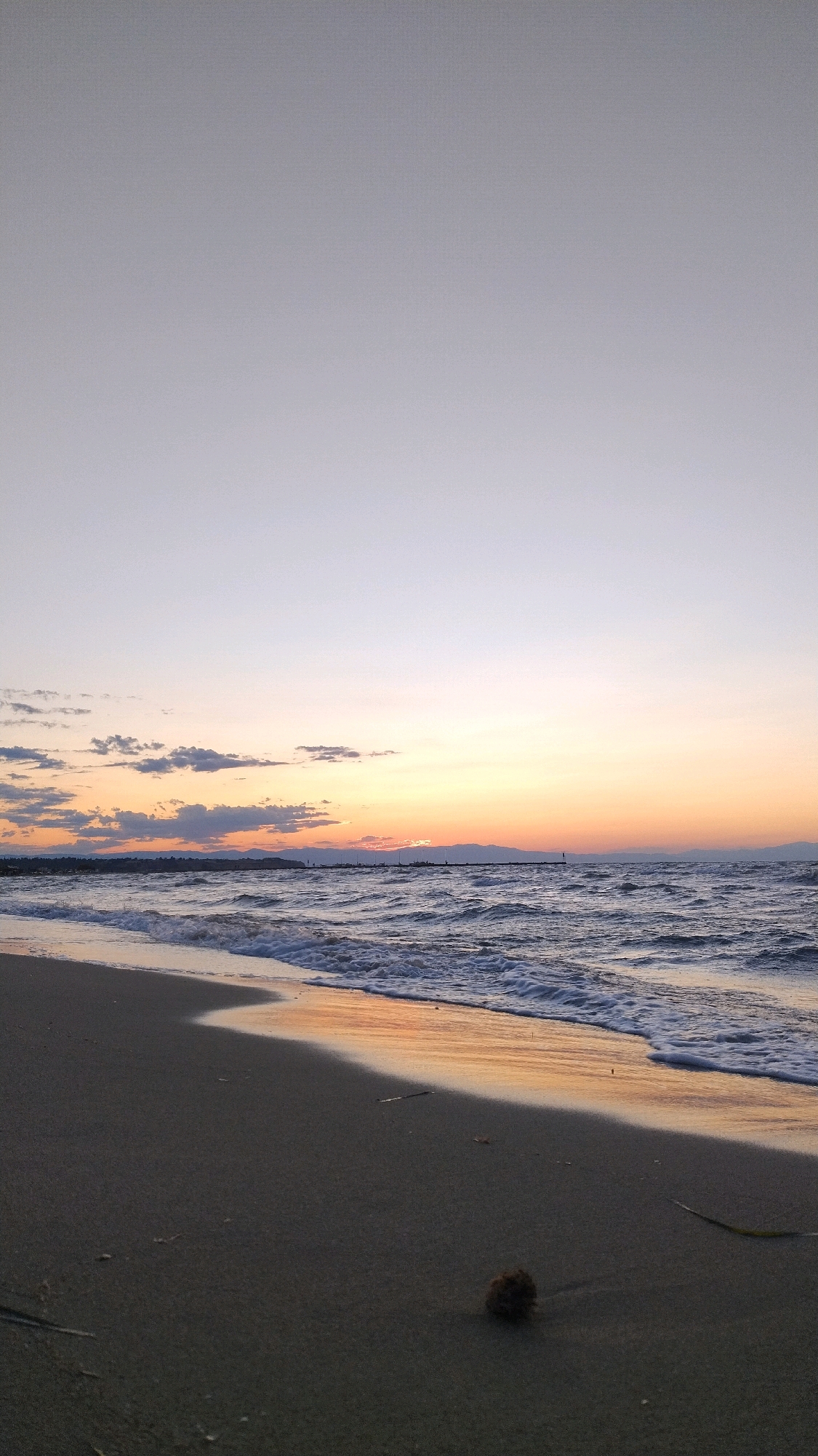 This is a vertical, low-angle shot of a beach at dusk in Neoi Epivates, Greece. The scene is dominated by a vast, gradient sky transitioning from a pale gray at the top to hues of soft orange and yellow near the horizon. Wispy clouds are scattered across the sky. The sea is choppy with moderate waves crashing onto the shore, leaving a line of white foam at the water's edge. The wet sand reflects the warm colors of the sunset, creating a shimmering effect along the shoreline. The beach itself is composed of fine, dark sand with some scattered debris, including a small, dark, spherical object in the foreground. In the distance, a dark landmass with a silhouetted coastline can be seen, possibly mountains, with a thin line of structures and a distant lighthouse or beacon visible. The overall atmosphere is tranquil and serene. There are no people or animals visible in the frame.