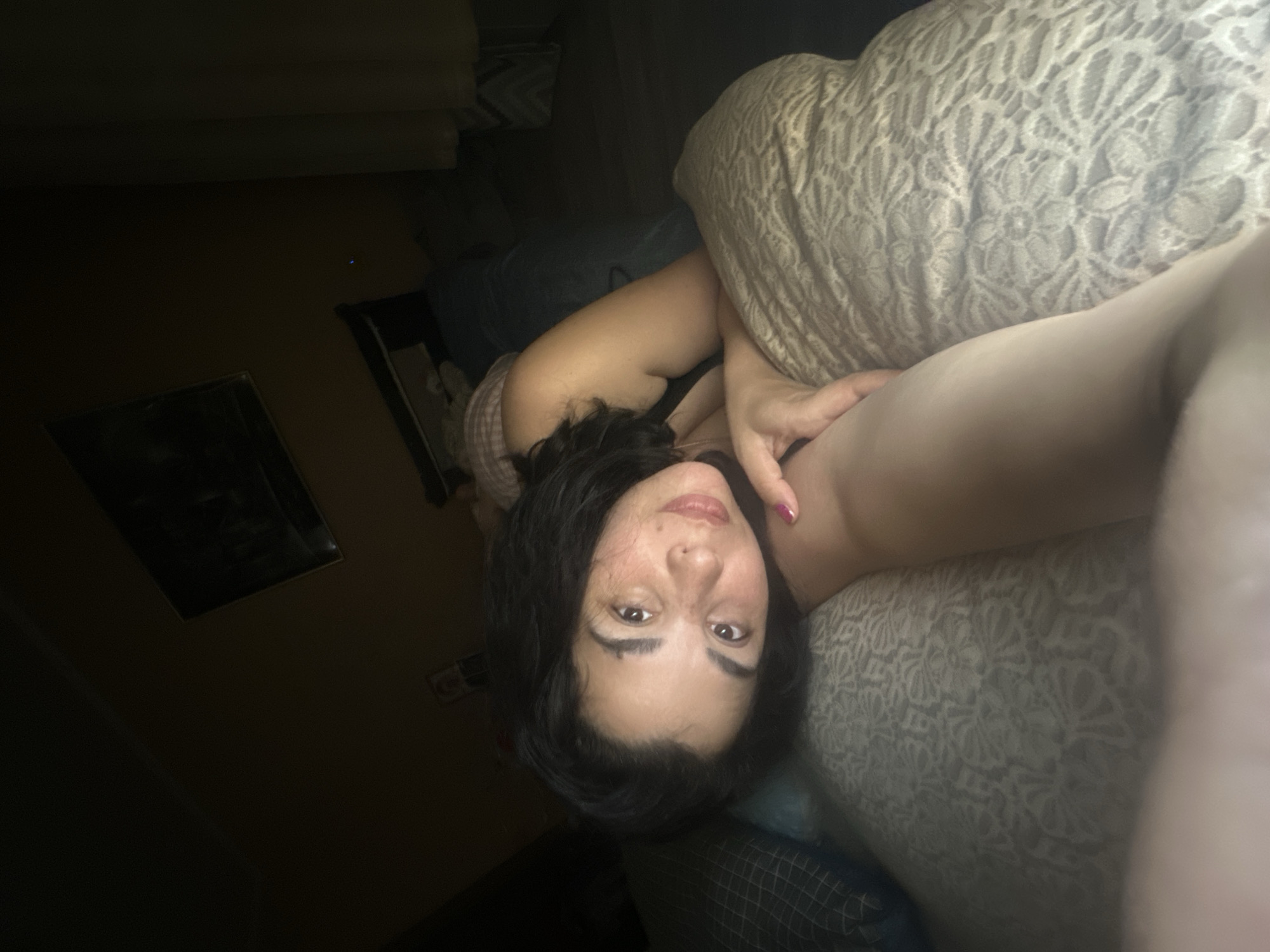 In an indoor, dimly lit bedroom, a young woman with dark, wavy hair is lying on a bed. She is positioned on her left side, with her head resting on a patterned pillow. Her face is tilted towards the camera, and she is looking directly at it with a slight smile. She is wearing a dark-colored bra. Her left arm is bent at the elbow, and her hand rests on the pillow near her face. The background is dark, but a framed picture is visible on the wall above her. To the right of the woman, a curtain can be seen. The scene appears to be a selfie taken in a private space, likely a bedroom.
