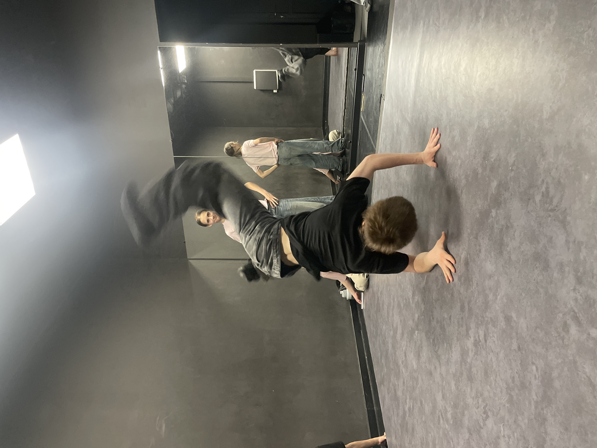 A photograph taken in a dance studio in Krasnoyarsk, Russia, shows a young man performing a dance move. He is upside down, with his hands on the floor, wearing a black shirt and gray pants. His legs are in the air, blurred from movement. In the background, a mirror reflects the scene, showing another person standing with their hands on their hips, wearing a pink shirt and jeans. The studio has a dark gray wall and a lighter gray floor. A square light fixture is visible on the wall.