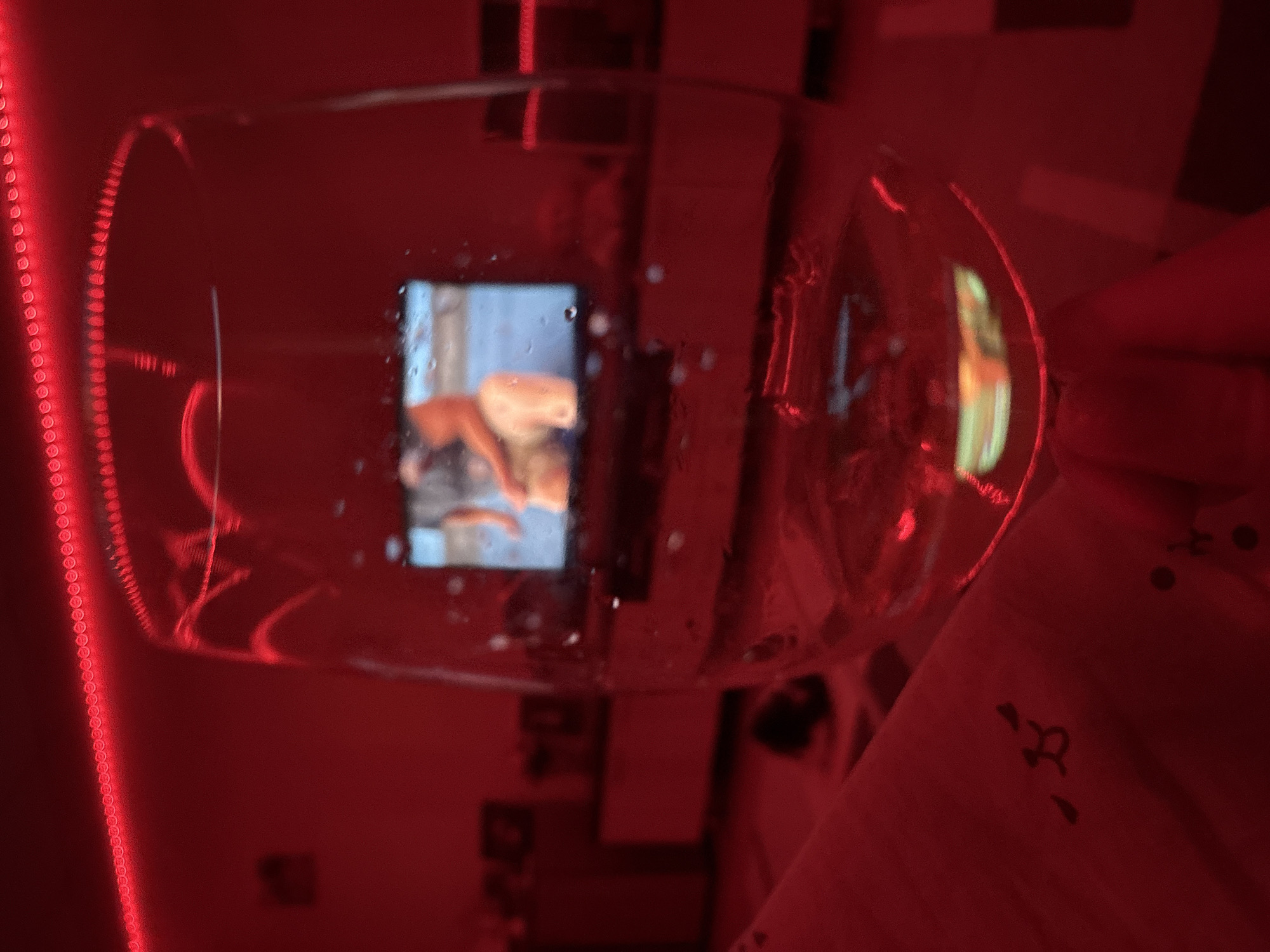 The image shows a wine glass in a room lit with red light, possibly from an LED strip. Inside the glass, a reflection of a phone screen displays an image of a person. The background is blurry, but suggests a bedroom environment with furniture and bedding. The location, Ust-Ilimsk, Russia, cannot be confirmed or denied based on the visual information alone.