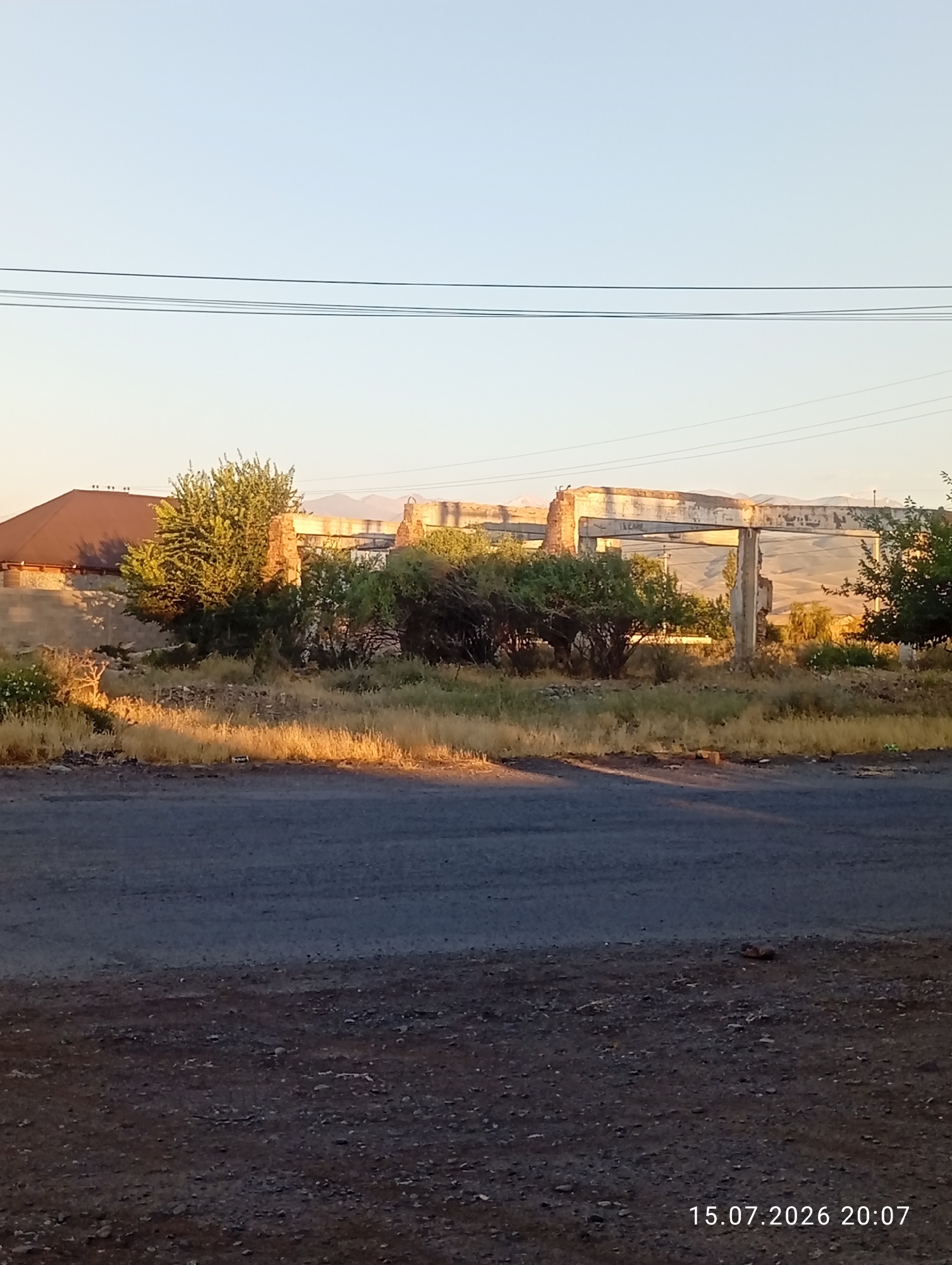The image shows a derelict concrete structure and surrounding overgrown vegetation in an outdoor setting. To the left, a building with a brown roof is partially visible, along with a stone wall. In the center and right, the remains of a large, open-air concrete structure are evident, with concrete pillars and beams in disrepair. Several trees and bushes are scattered throughout the scene, some of which appear to be dead or withered, particularly in the foreground where dry grass and debris are present. Power lines are visible stretching across the top of the frame against a clear sky. In the background, distant hills or mountains can be seen. The ground in the foreground is a rough, dark asphalt or dirt surface. A timestamp in the lower right corner indicates "15.07.2026 20:07". The scene appears to be located in the city of Ak-Suu, Kyrgyzstan.