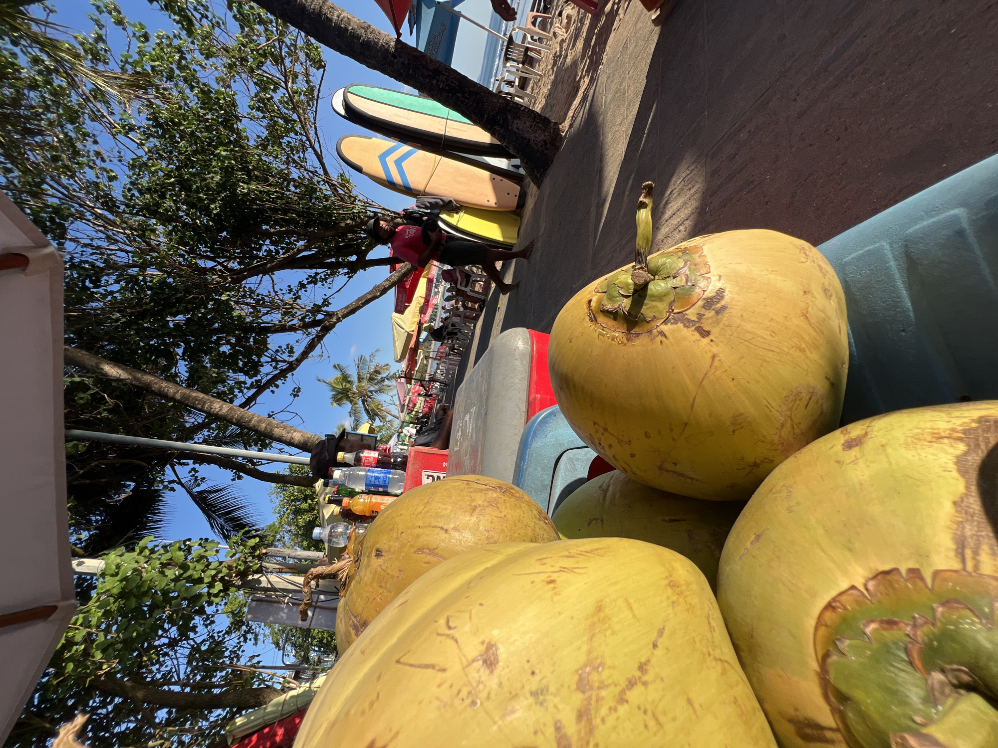 In Kuta, Indonesia, an outdoor vendor displays a cart filled with large, yellow coconuts in the foreground. Behind the coconuts, a table is set up with various bottles of drinks, including water, orange soda, and blue liquid. A person in a pink shirt and black shorts, wearing a hat, stands near a collection of surfboards leaning against a tree, holding a camera or bag. The background shows a beach scene with umbrellas, tables, and chairs, and the ocean with gentle waves. The sun is shining, casting shadows on the paved walkway.