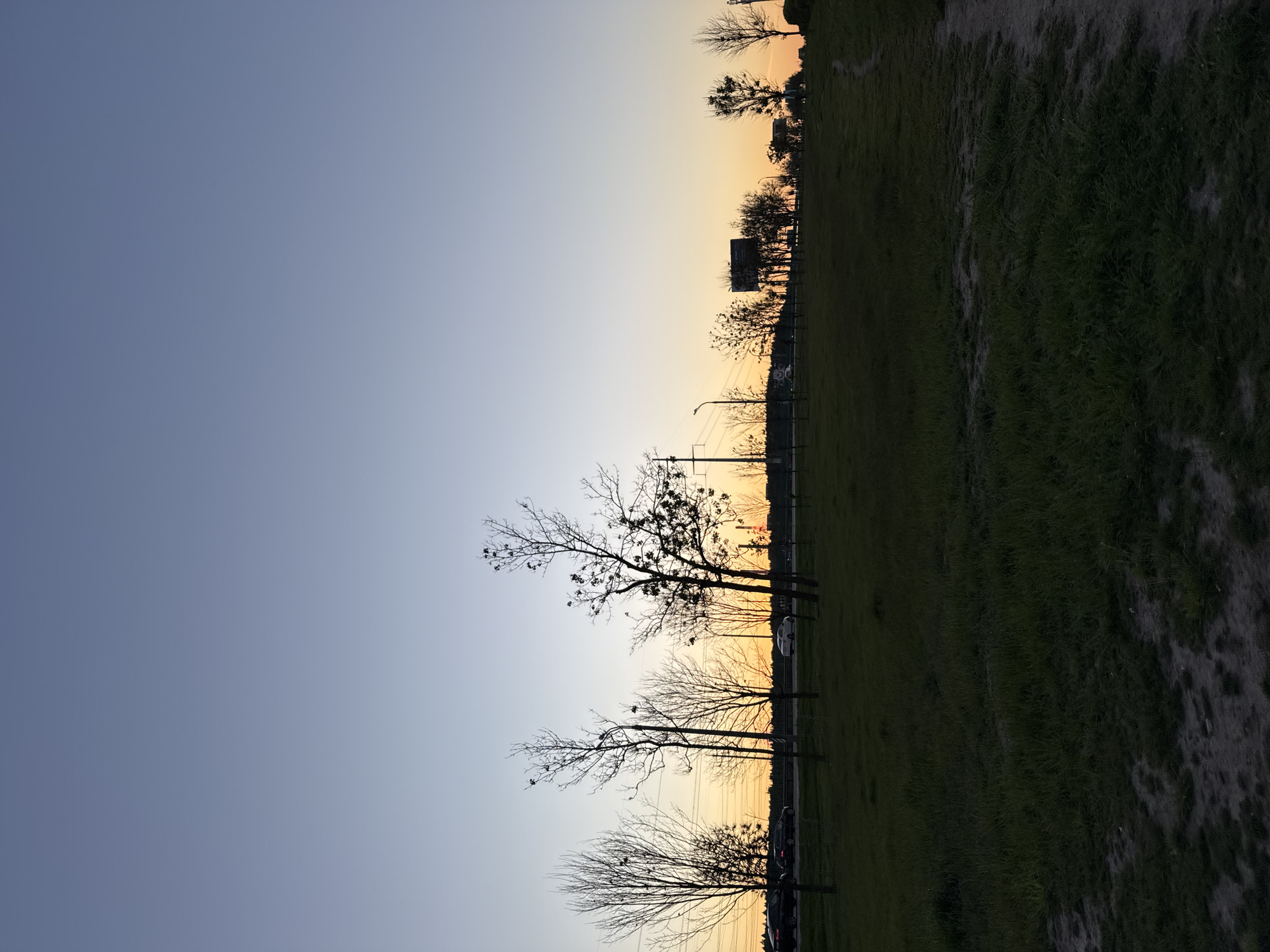 The image shows a park landscape during sunset in Minsk, Belarus. The sky transitions from blue to orange. Silhouetted trees line the horizon. A grassy area stretches across the bottom half of the frame. Utility poles and some buildings are visible. Cars are parked along a road adjacent to the grassy area. There are no visible people.