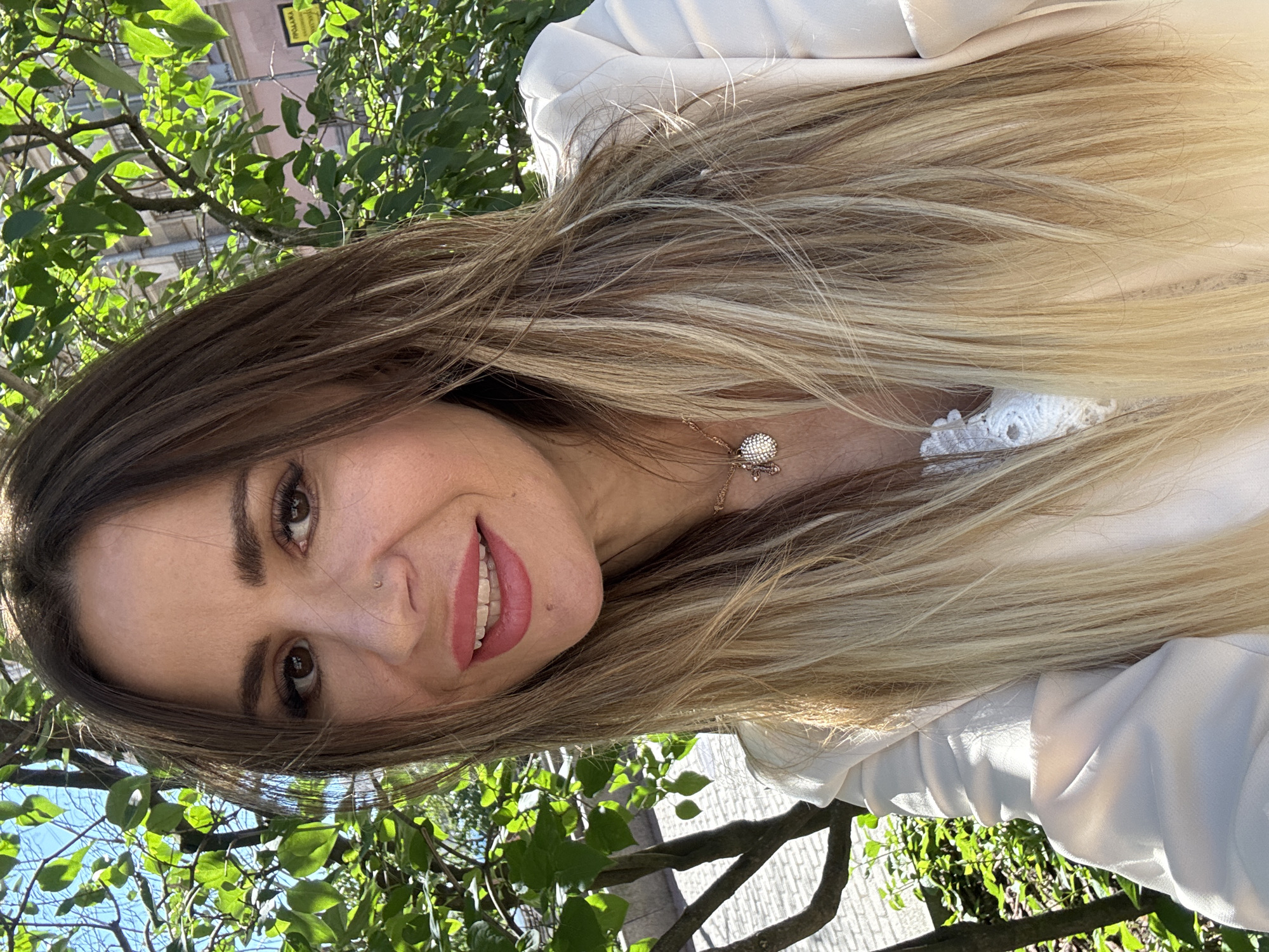 A woman is captured in a selfie taken outdoors. She has long, layered hair that transitions from dark brown to blonde. Her eyes are brown, and she is wearing makeup, including pink lipstick and mascara. She has a small nose stud. She is wearing a light-colored blazer over a white lace top and a necklace with a pendant. The background suggests an urban setting in Saint Petersburg, Russia, with green foliage, trees, and buildings visible. The sunlight indicates it is daytime. The texture of the paving stones in the background suggests she might be in a pedestrian area or a park. A yellow sign with Cyrillic text is partially visible on a building in the background.