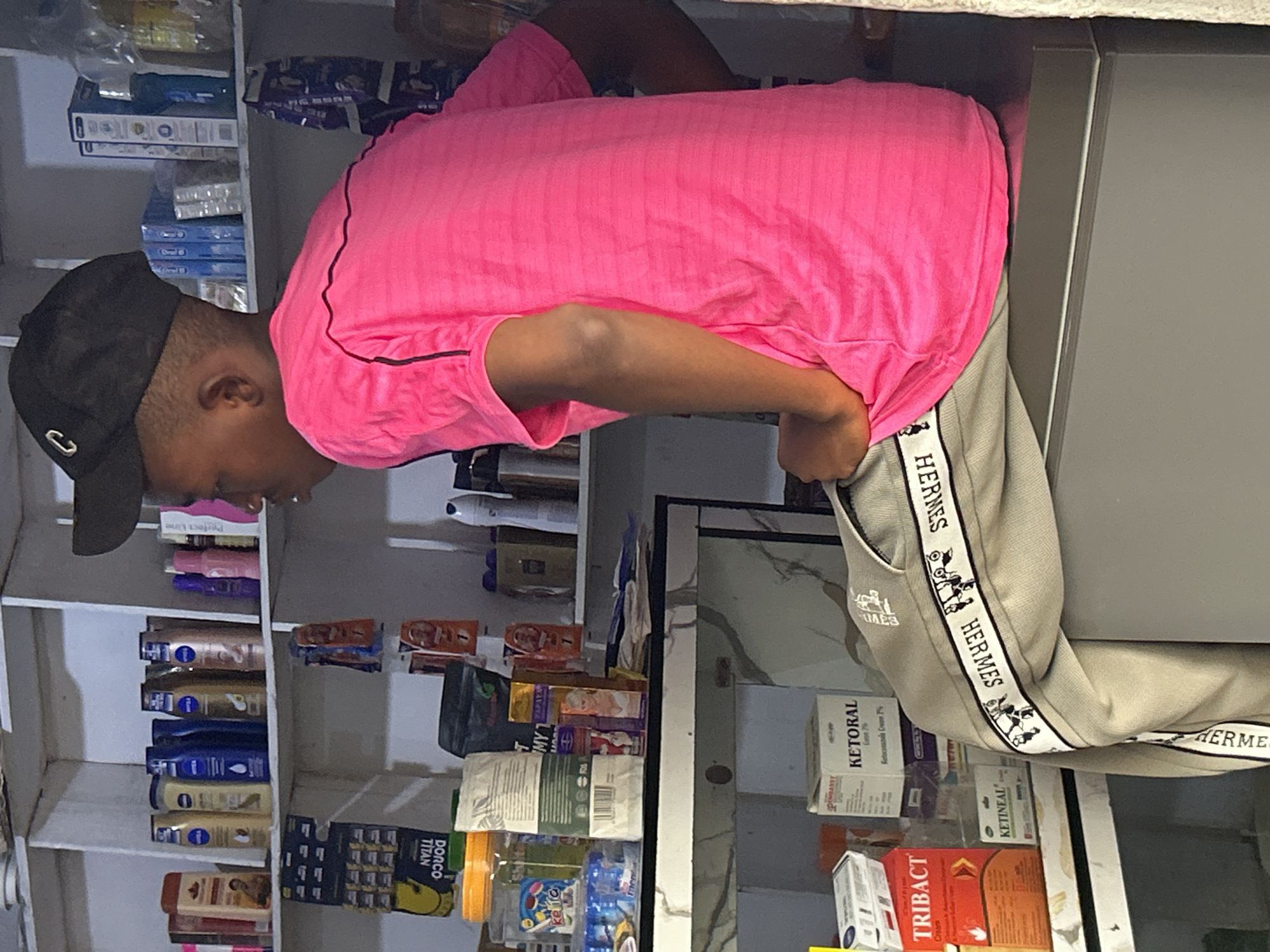 The image depicts the interior of a small shop, likely a pharmacy or convenience store, located in Ihiagwa, Nigeria. A young person wearing a bright pink t-shirt and grey pants with a distinctive "HERMES" logo on the side is seated on a counter. The shelves behind them are stocked with various products, including toiletries, medicines, and personal care items. Some of the visible brands include NIVEA, TRIB ACT, and KETORAL. The setting appears to be dimly lit, with artificial lighting illuminating the shelves. The overall impression is that of a bustling local retail environment.