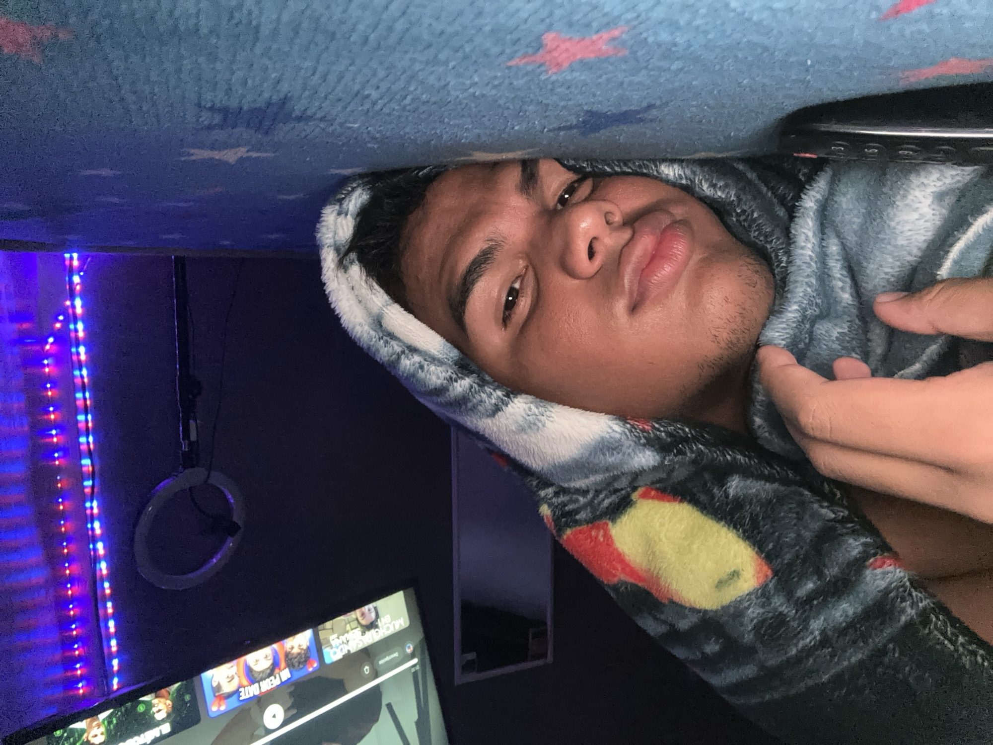 A person is lying beneath a blue blanket with star patterns. The person has dark hair, brown eyes, and appears to be male. He is partially wrapped in a grey and black blanket with yellow and red patches. In the background, there is a television screen displaying various content, a ring light hanging on the wall, and LED strip lights emitting blue and red light. The location, Parroquia Ocumare, Venezuela, cannot be verified from the image.
