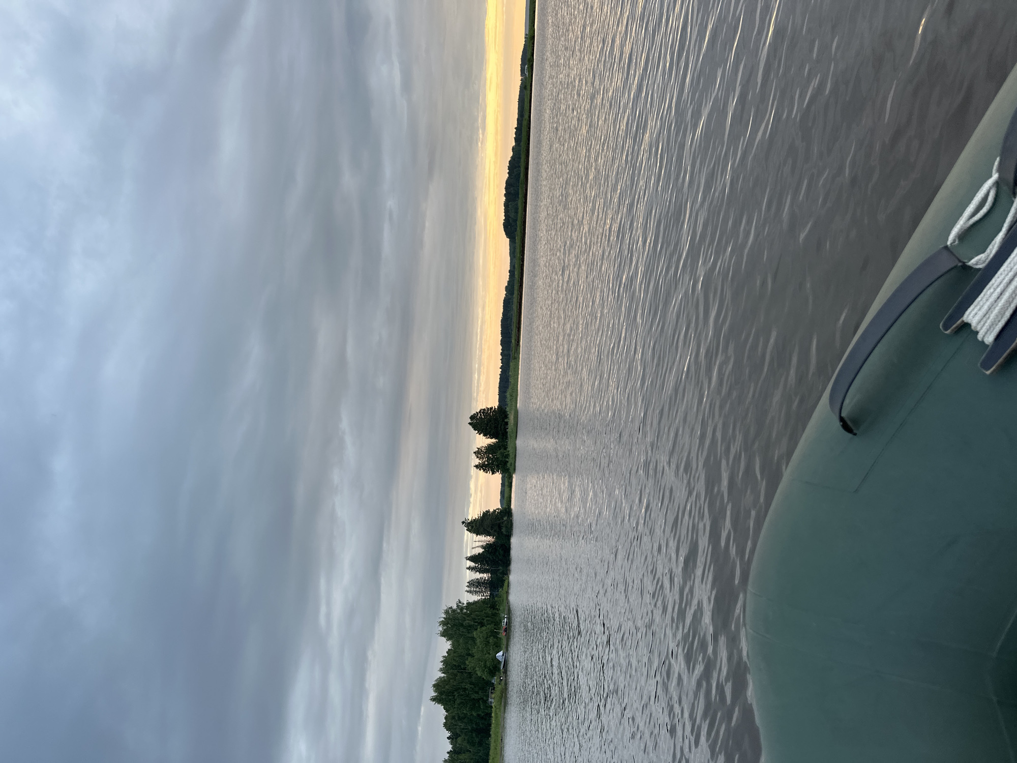 An over-the-shoulder shot from a boat shows a calm lake with a distant tree-lined shore under a cloudy sky. In the foreground, the green inflatable boat has a rope coiled on its deck. The lake water is dark and rippled. The far shore is mostly covered in dense green trees, with a few smaller islands also visible. The sky is overcast with grey clouds, but a sliver of yellow light on the horizon suggests the sun may be setting or rising. The location is Shul'dikha, Russia.