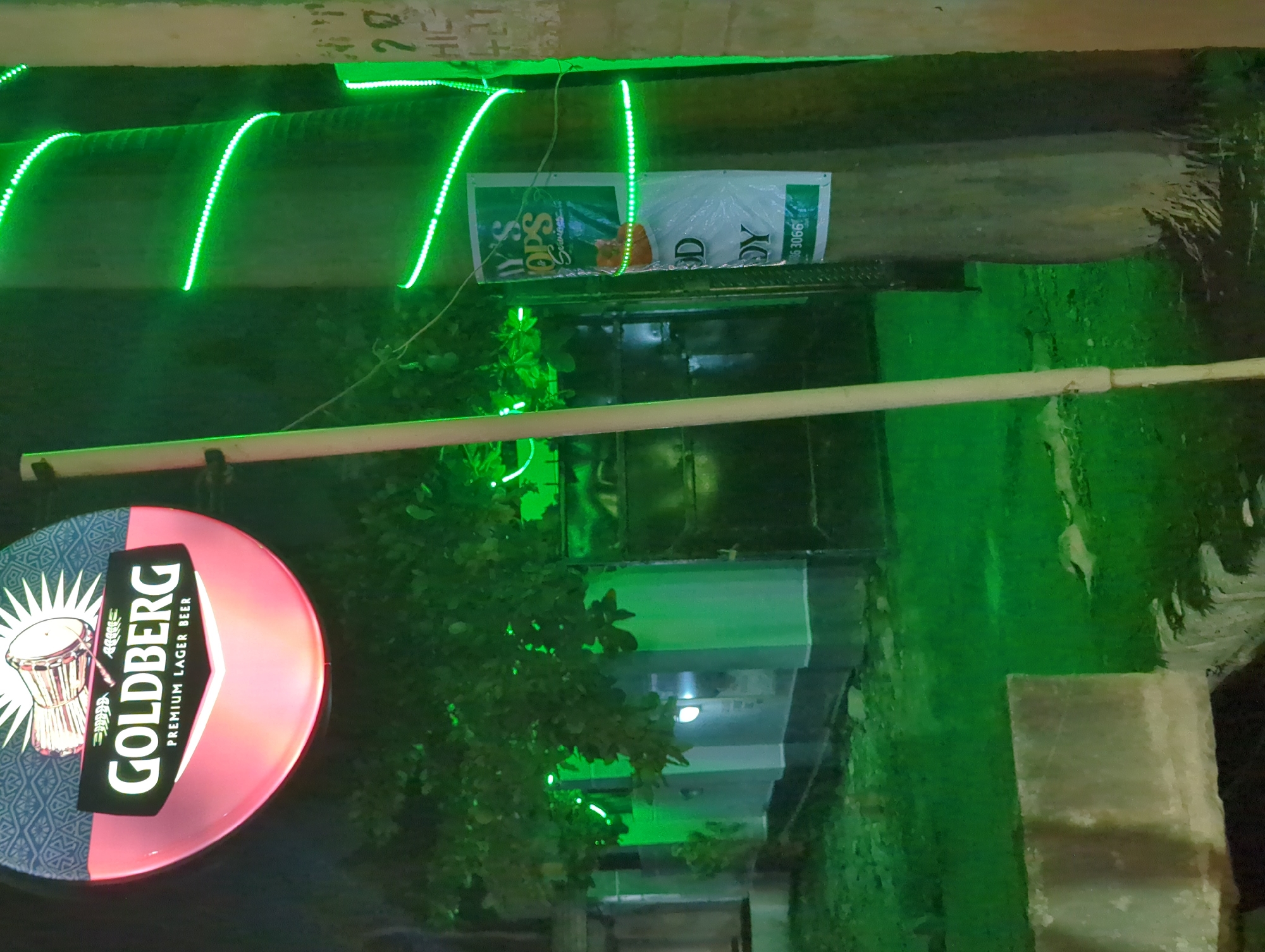 The image shows a nighttime scene in Narayi, Nigeria. A "Goldberg Premium Lager Beer" sign is visible on the left. Green LED lights are strung along the upper portion of a building facade. A "DY TOPS" sign is above a dark doorway. Vegetation, likely a small tree, obscures part of the view. The environment has an overall green hue from the lighting.