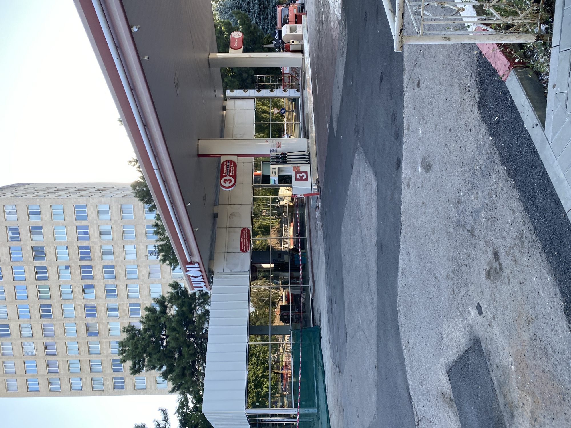 The image shows a Lukoil gas station, likely in Chișinău, Moldova, with a tall building in the background. The gas station appears to be undergoing renovations, with construction barriers and materials visible. Several fuel pumps are present, and a sign indicates "Premium-95 Motorina." A red truck is parked near the service area. The overall scene suggests a commercial area with ongoing maintenance or upgrades.
