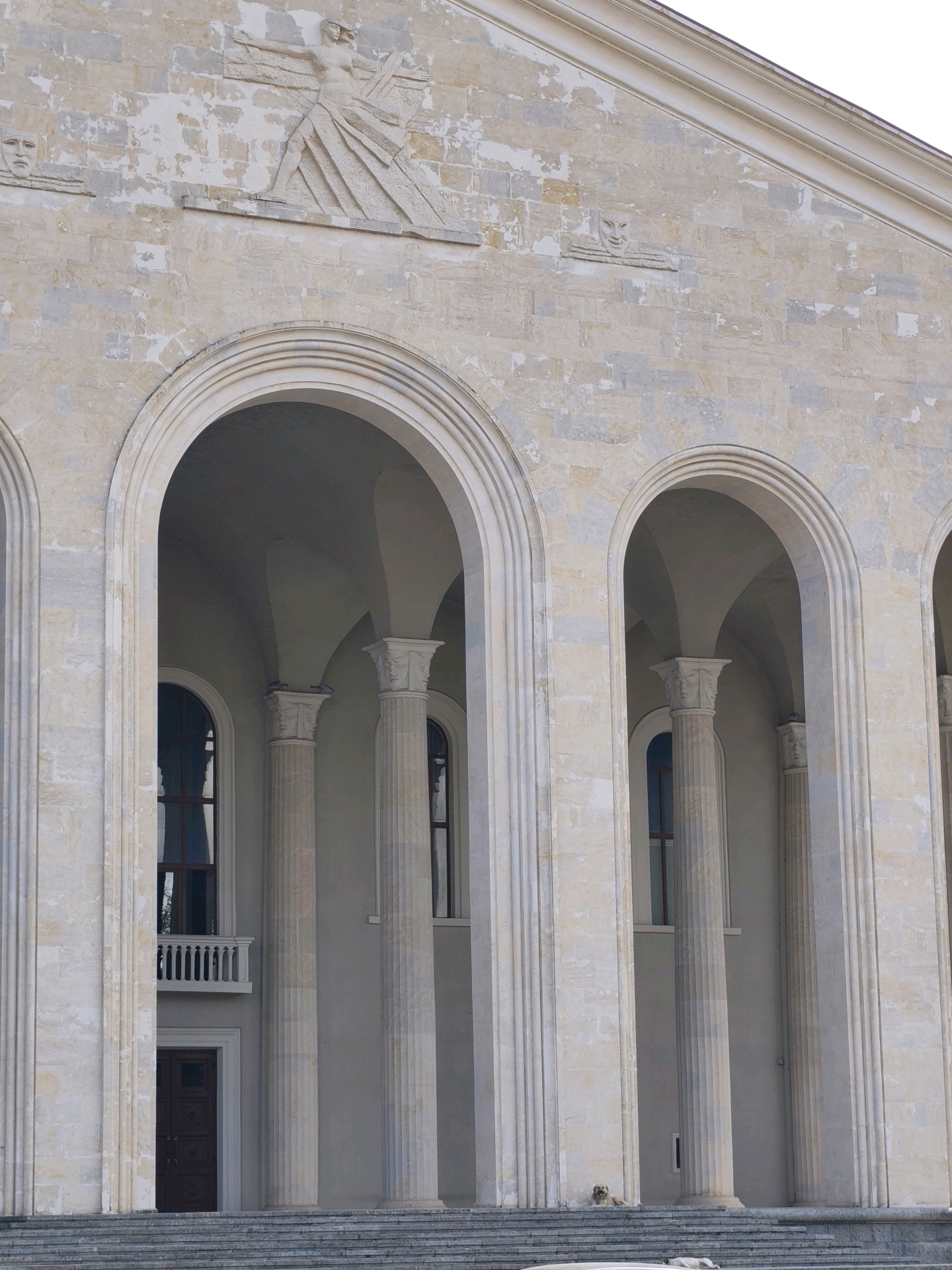 The image shows a detailed view of the exterior of the Ozurgeti Drama Theatre in Ozurgeti, Georgia. The building features a grand facade with a series of large, arched openings supported by fluted columns. Above the arches, the stone facade is adorned with bas-relief sculptures. Notably, there is a large triangular frieze with a central figure of a person with outstretched arms, and two smaller masks on either side. The stonework shows signs of weathering and some paint peeling. Within the arched openings, tall, narrow windows are visible, some with a small balcony. A dark-colored double door is also visible at ground level, accessed by a wide set of stone stairs. The overall impression is of a monumental and somewhat aged public building.