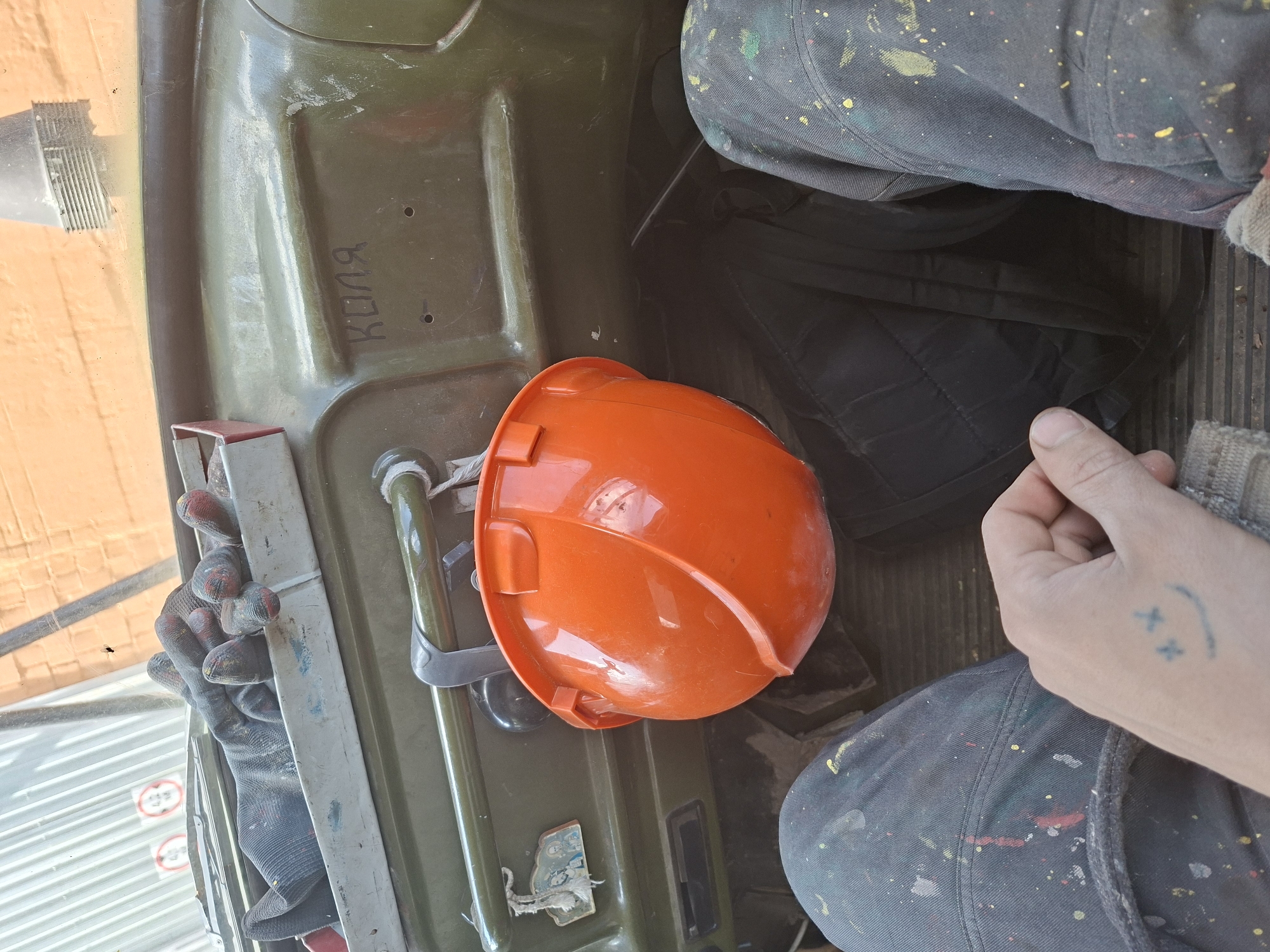 The scene appears to be inside a vehicle, possibly a truck or heavy machinery, as indicated by the olive green, heavy-duty interior. An orange hard hat rests on a section of the interior. A pair of work gloves is visible. A person is present; portions of their paint-splattered pants and hand are visible. The hand has a small tattoo resembling a smiley face with "X" eyes. The location is unknown, and there is no information to confirm the location is Zhabinka, Belarus.