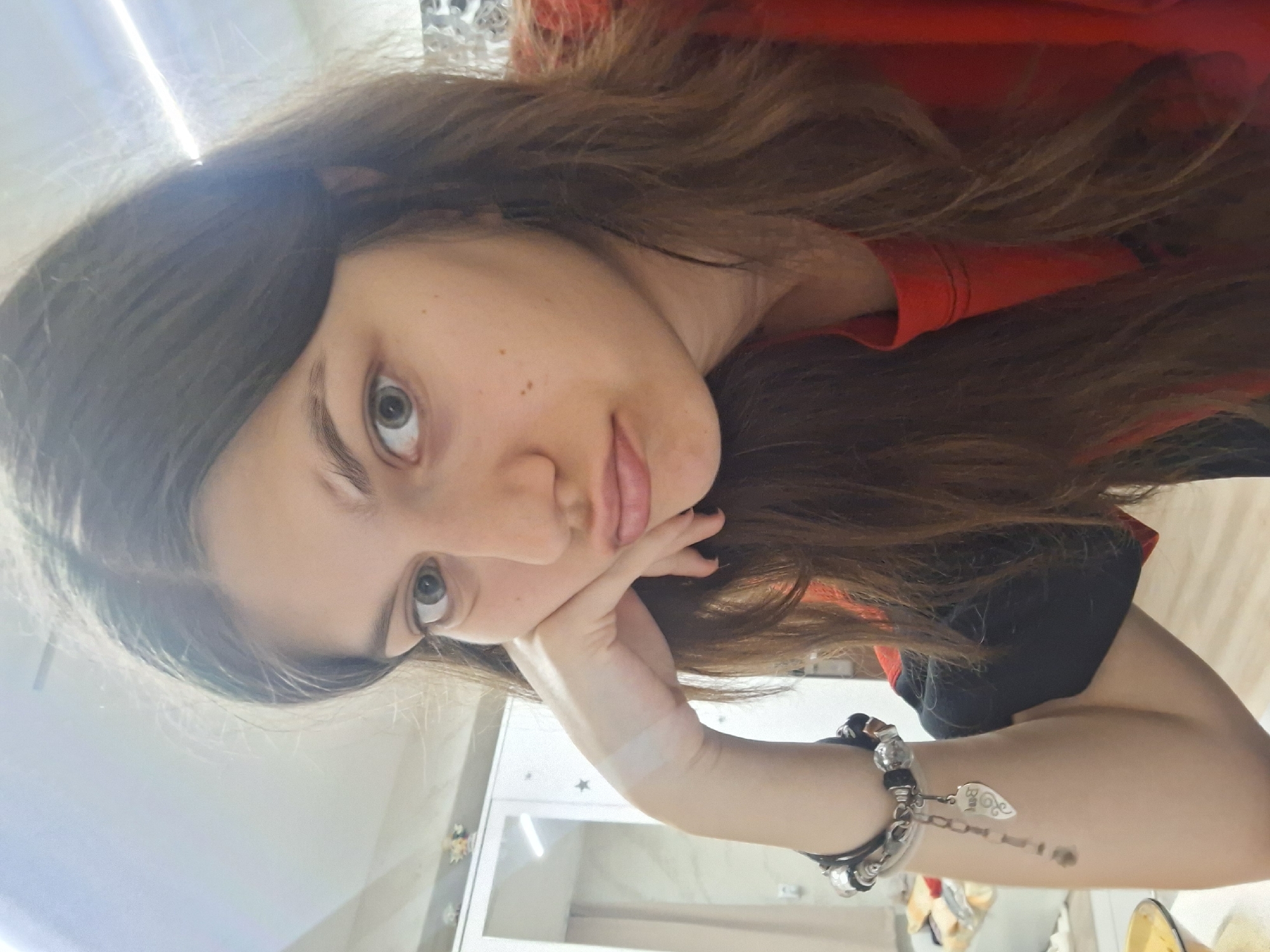 A young woman with long brown hair is posing for a selfie. She has fair skin, light eyes, and is wearing a red and black top. Her right arm is bent, with her hand resting on her cheek. On her wrist is a multi-beaded bracelet with a heart-shaped charm. The background is slightly out of focus, but hints at a modern interior, possibly a room in a house. The location in the city of Panagyurishte, Bulgaria is not identifiable from the image.