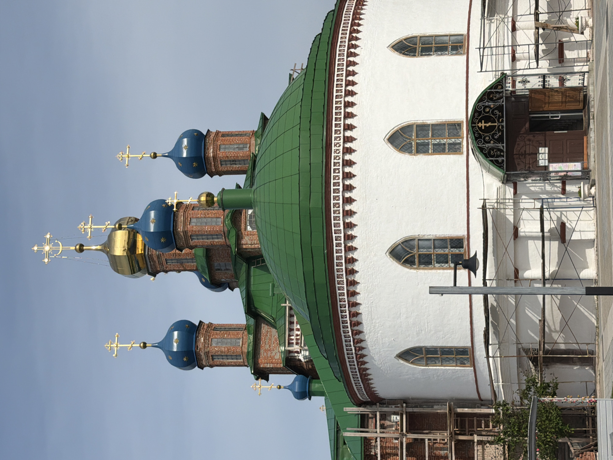 The image captures a detailed view of the Church of the Transfiguration in Usolye, Russia, on a clear day. The church features a prominent, large, green-tiled dome at the forefront, with a circular white base adorned with a decorative cornice and arched windows. Above the main dome, several smaller, blue-domed towers with golden crosses rise, interspersed with red brickwork and a central golden dome. Scaffolding is visible on the left side of the building, indicating ongoing construction or renovation. A dark arched entrance with ornate metalwork and a golden cross is centrally positioned at the base of the church. To the left of the entrance, a lamppost stands, and a small tree with some foliage is visible near the scaffolding. The ground in front of the church appears to be paved. The sky above is a pale blue with slight cloud cover. No people are visible in the frame.