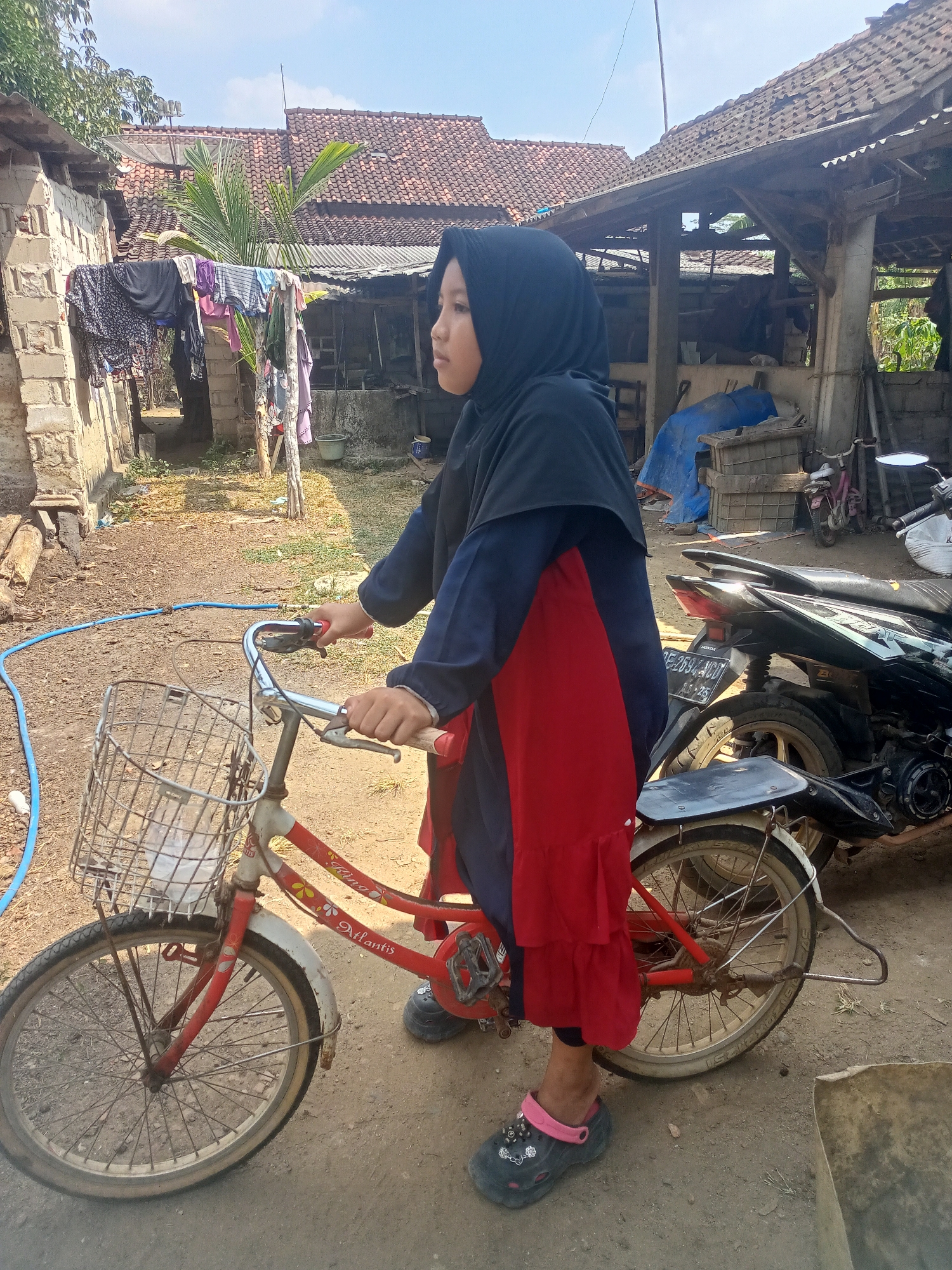 A young girl wearing a black hijab and a navy blue and red dress stands next to a red bicycle with a wire basket in front. She is looking to her right and holding the handlebars. Her left foot is visible, wearing a dark gray clog with a pink strap. The bicycle has a white front fender and a red frame with floral decals. The ground is dirt and some gravel. In the background, there are several structures with tiled roofs, some appearing to be houses and a partially open shelter. Clothes hang on a line to the left of the girl. A black motorcycle is parked behind the bicycle. The scene appears to be in a rural or semi-rural area, possibly a village. The overall lighting suggests a sunny day.