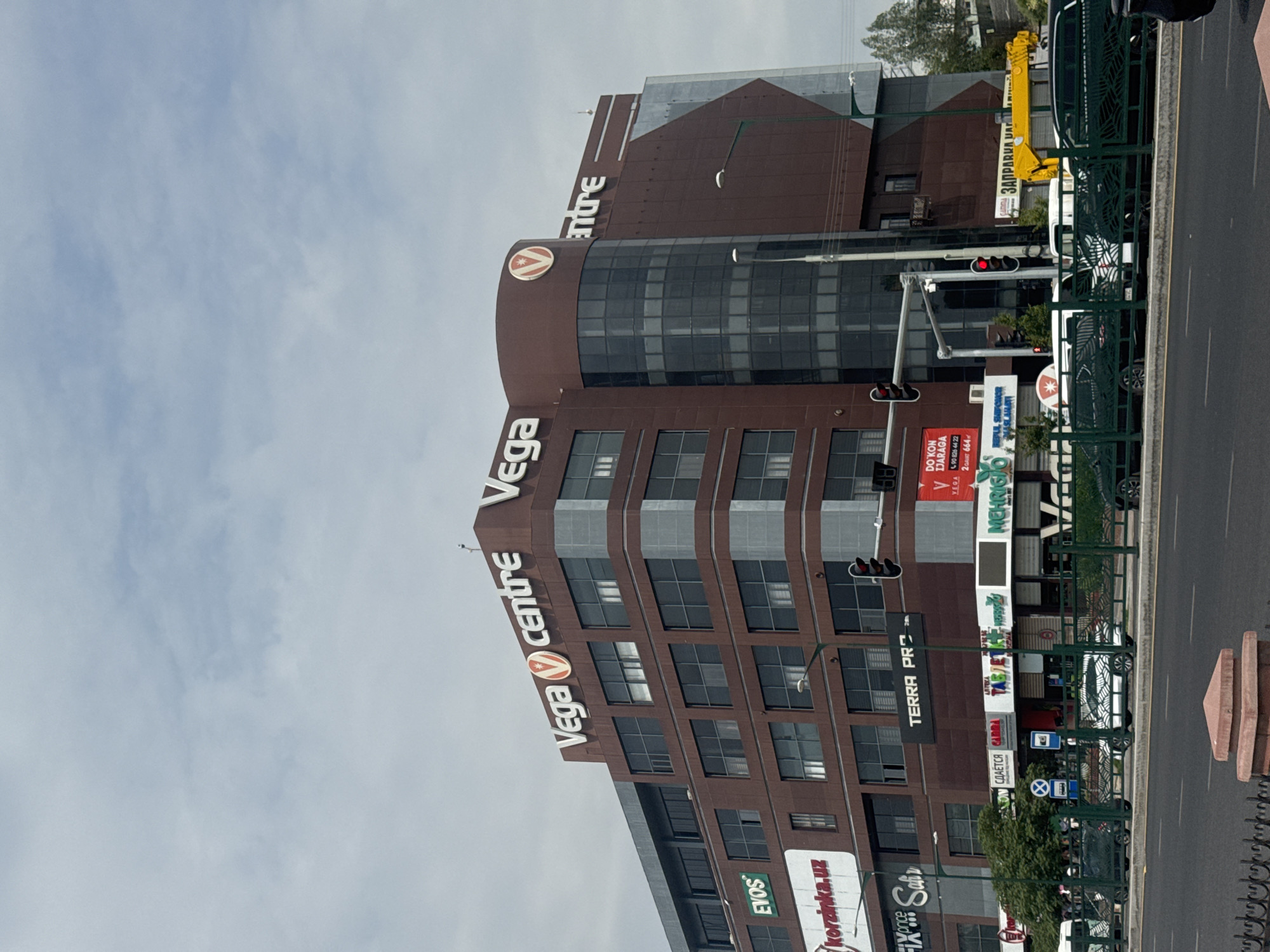 This is a frontal, eye-level medium shot of the Vega Centre, a modern, multi-story commercial building in Tashkent, Uzbekistan. The building is predominantly reddish-brown with large, dark windows. Prominent signage includes "Vega Centre" in white lettering on the upper facade, with a red circular logo. Other visible signs on the building include "EVOS," "korzinka.uz," "TERRA PRO," and various Cyrillic text. A curved glass section is visible on the right side of the building. In the foreground, a street with multiple lanes of traffic is present. Several cars, including a white car with a patterned wrap, are visible on the street and parked behind a green metal fence. Traffic lights are also visible. The sky is overcast. The overall scene depicts a busy urban environment.